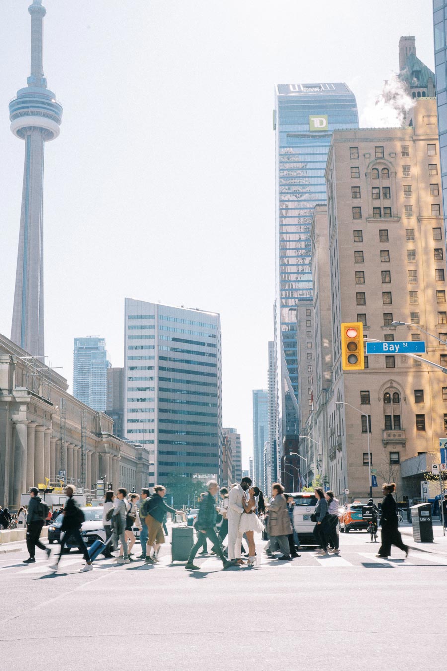 Cityscape of Toronto with the CN Tower, bustling pedestrians crossing a downtown intersection, surrounded by modern high-rise buildings under a clear sky.
