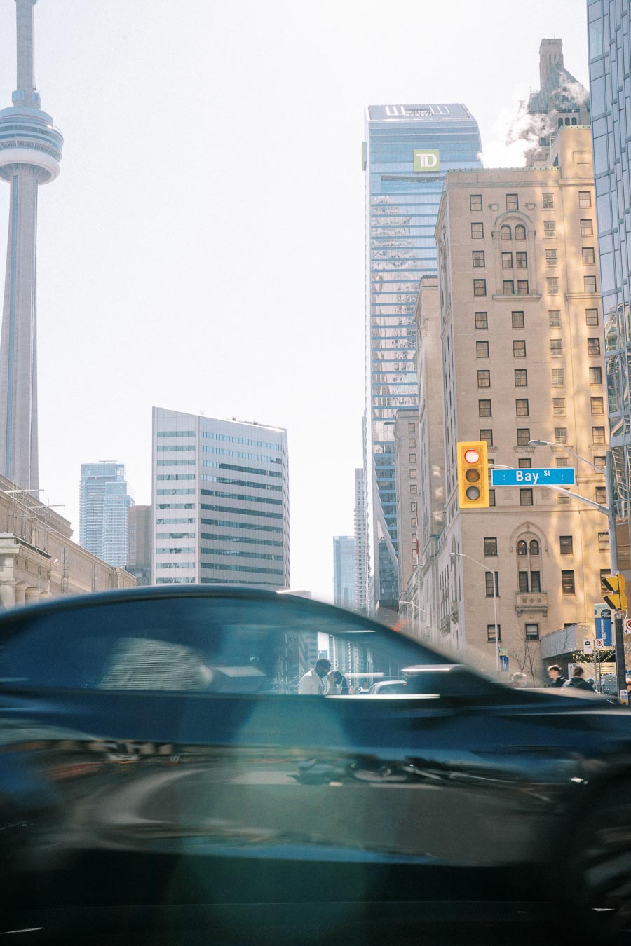 Urban city scene in Toronto with CN Tower in the background, busy street with a blurred car in the foreground, and towering skyscrapers on a bright day.