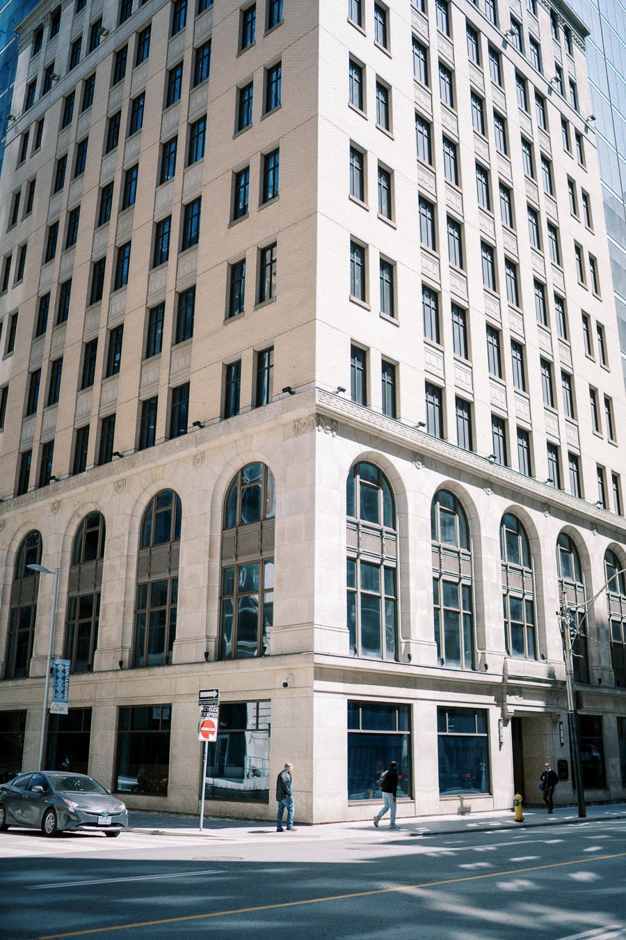 Urban street view with tall beige brick building featuring arched windows, pedestrians walking on the sidewalk, and a car parked nearby.