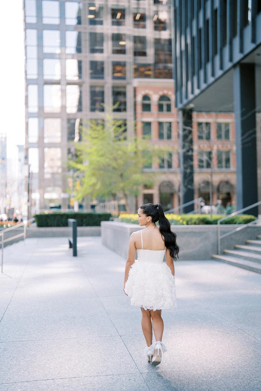 Woman in a white dress and heels walking on a modern city street, surrounded by tall buildings and greenery.