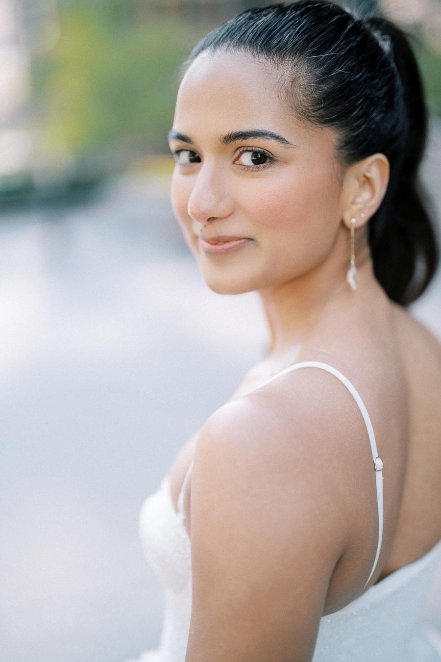 Young woman with a ponytail wearing a white dress and earrings, looking back with a slight smile, blurred outdoor background.
