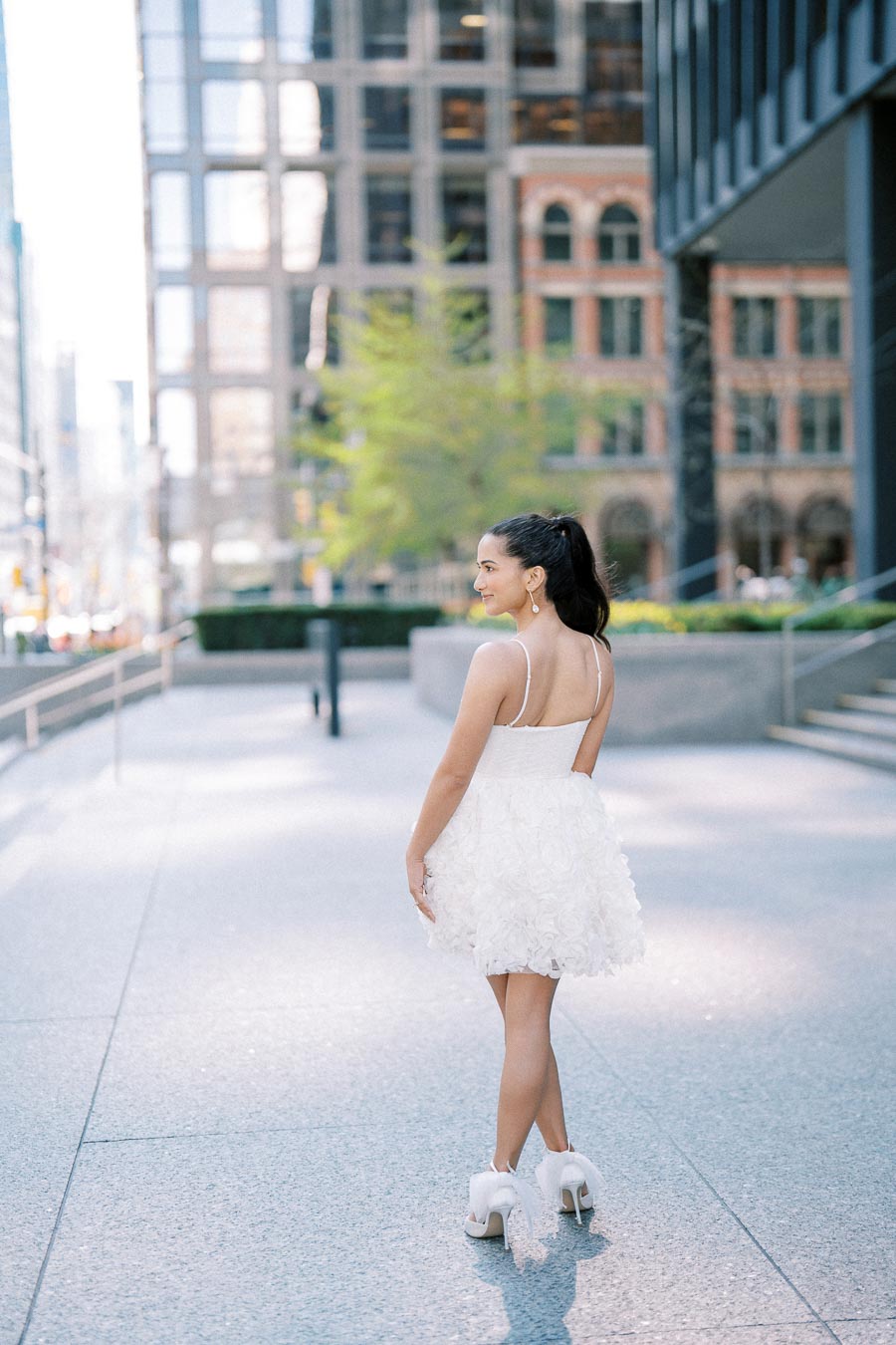 A woman in a short white dress and high heels walks down a city sidewalk, with modern buildings in the background.