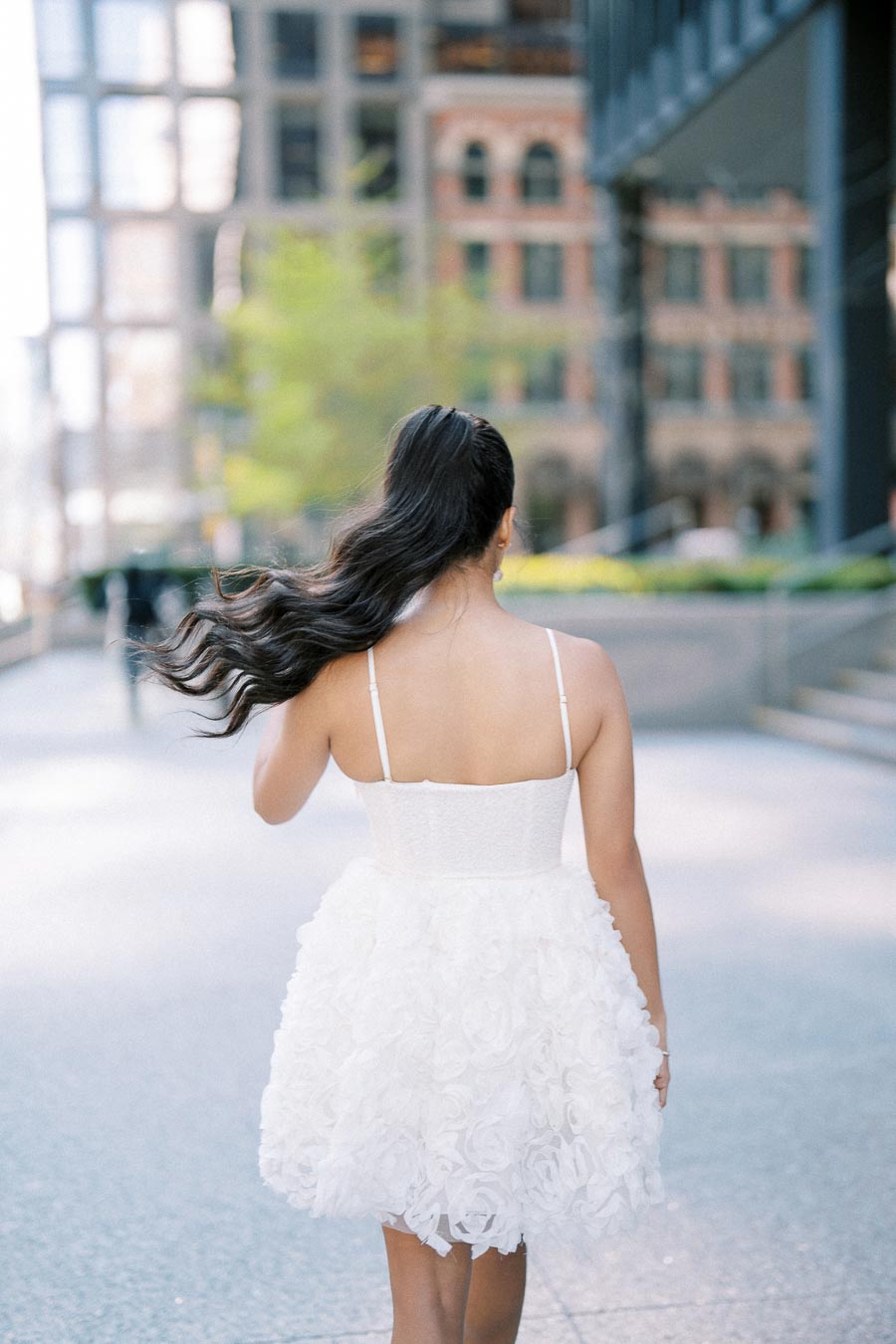 Woman in a white floral dress walking down an urban street with modern buildings in the background.