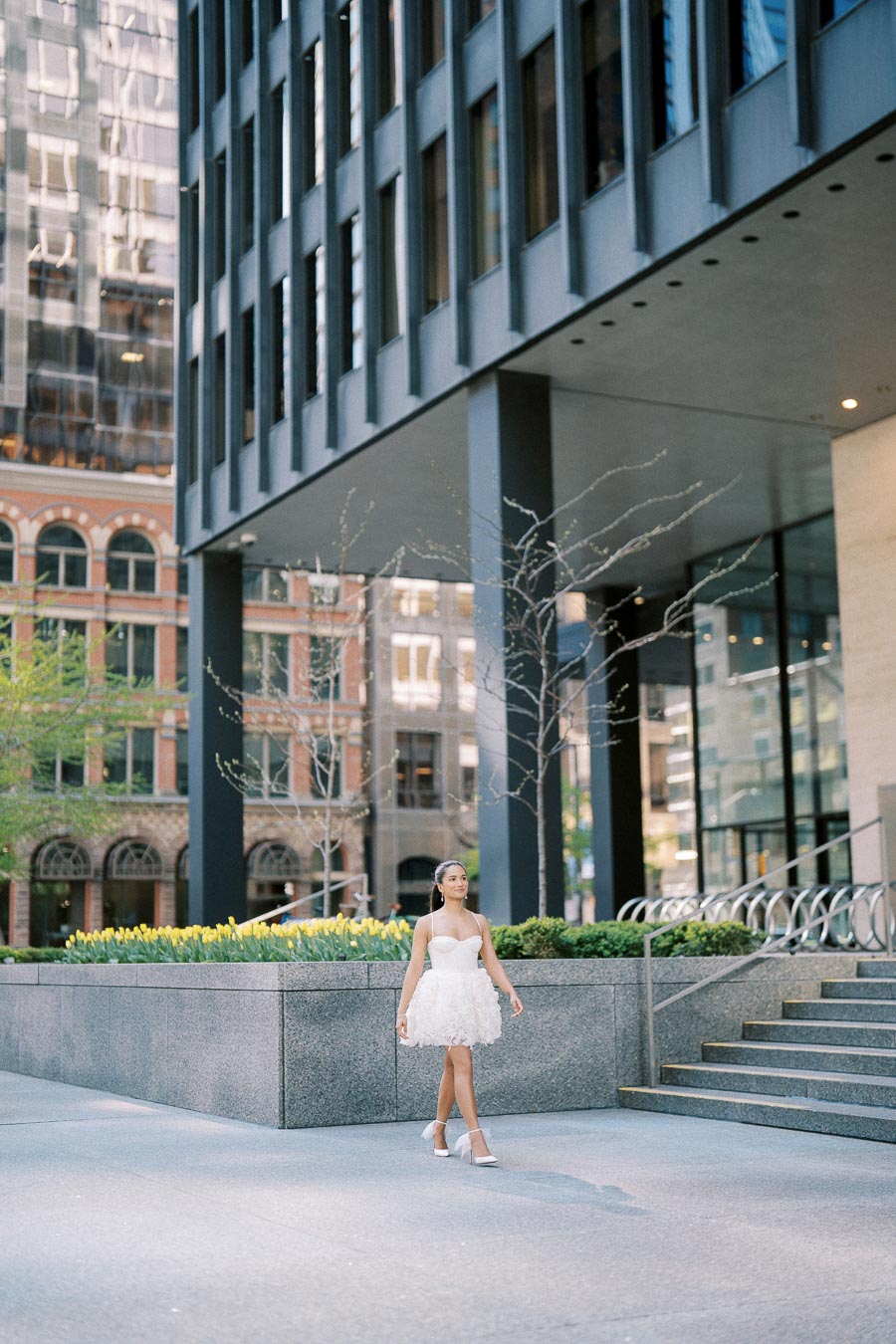 A woman in a white dress walking in an urban setting with modern architecture and yellow flowers in the background.