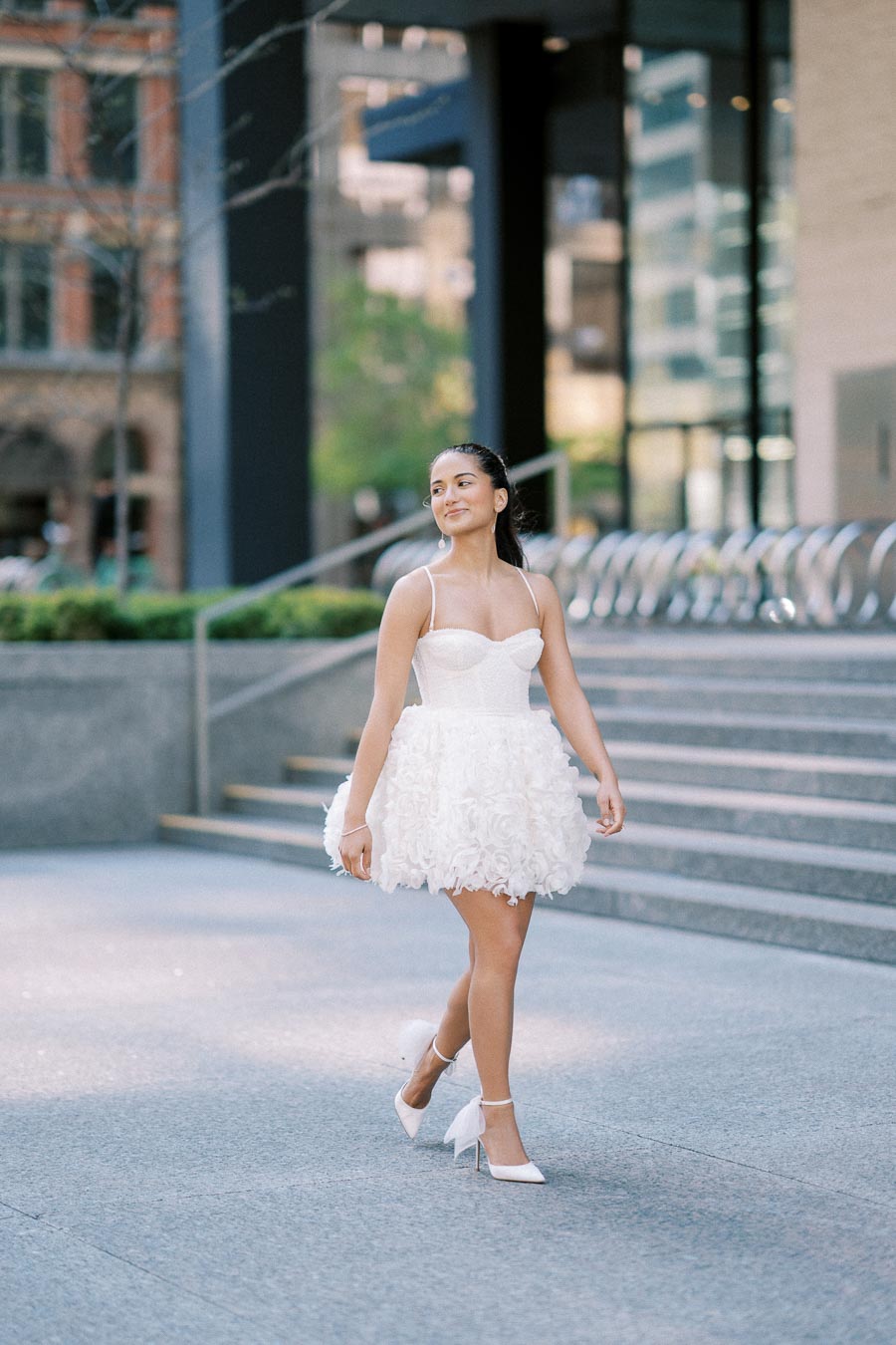 A person in a stylish white dress walking confidently on an urban sidewalk, steps and modern architecture in the background.