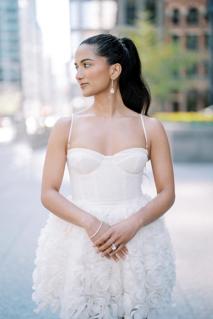 Young woman in an elegant white dress with floral details, standing outdoors in an urban setting, exuding a sophisticated and graceful look.