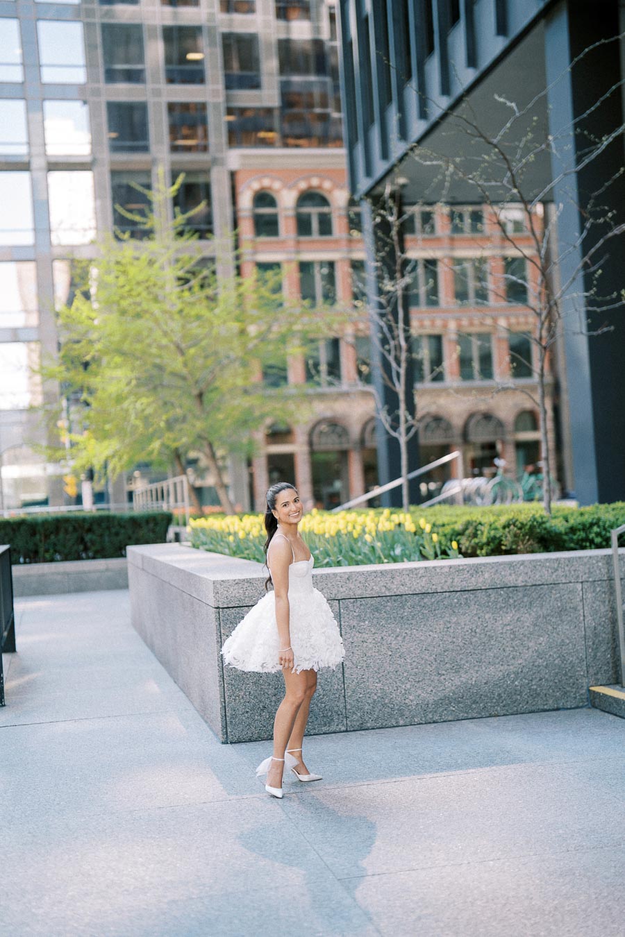 A woman in a white dress poses outdoors in an urban setting, surrounded by modern and historic buildings, with green plants and flowers in the background.