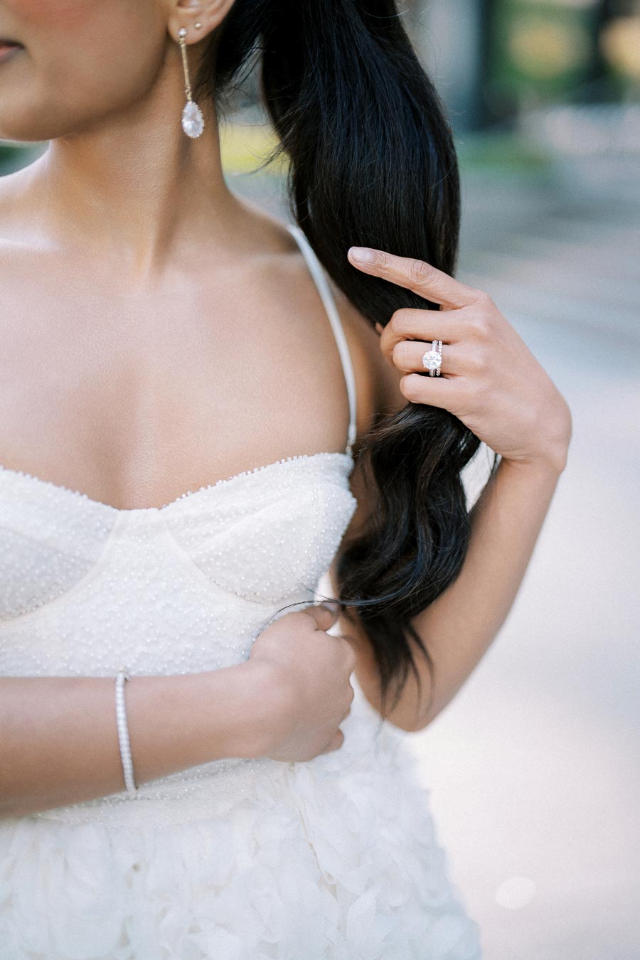 A bride in a white wedding dress, showcasing elegant jewelry including a diamond ring and drop earrings, while gently holding her long, dark hair.