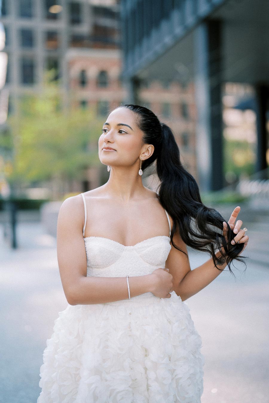 Young woman in an elegant white dress with floral details posing outdoors, holding her long dark hair with an urban background.