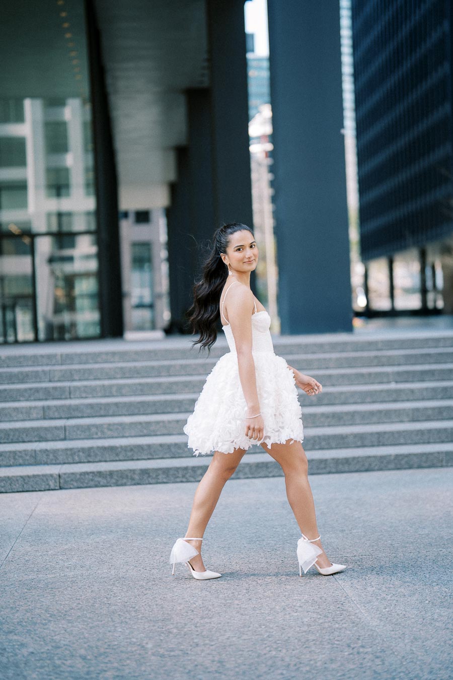 Young woman in a short white dress and heels walking confidently in an urban setting, showcasing chic fashion against a backdrop of modern architecture and steps.