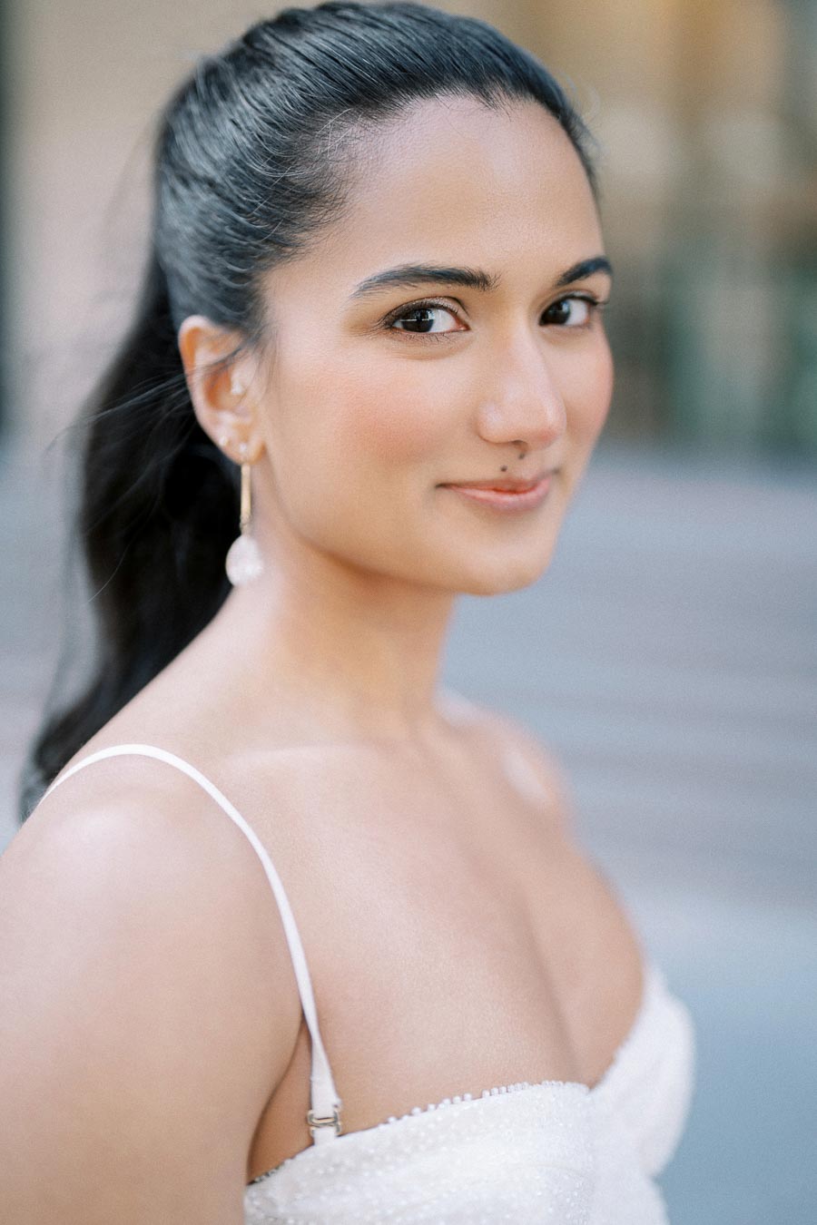 A woman with long dark hair in a ponytail, wearing a white dress and pearl earrings, looking at the camera with a soft smile, with a blurred urban background.