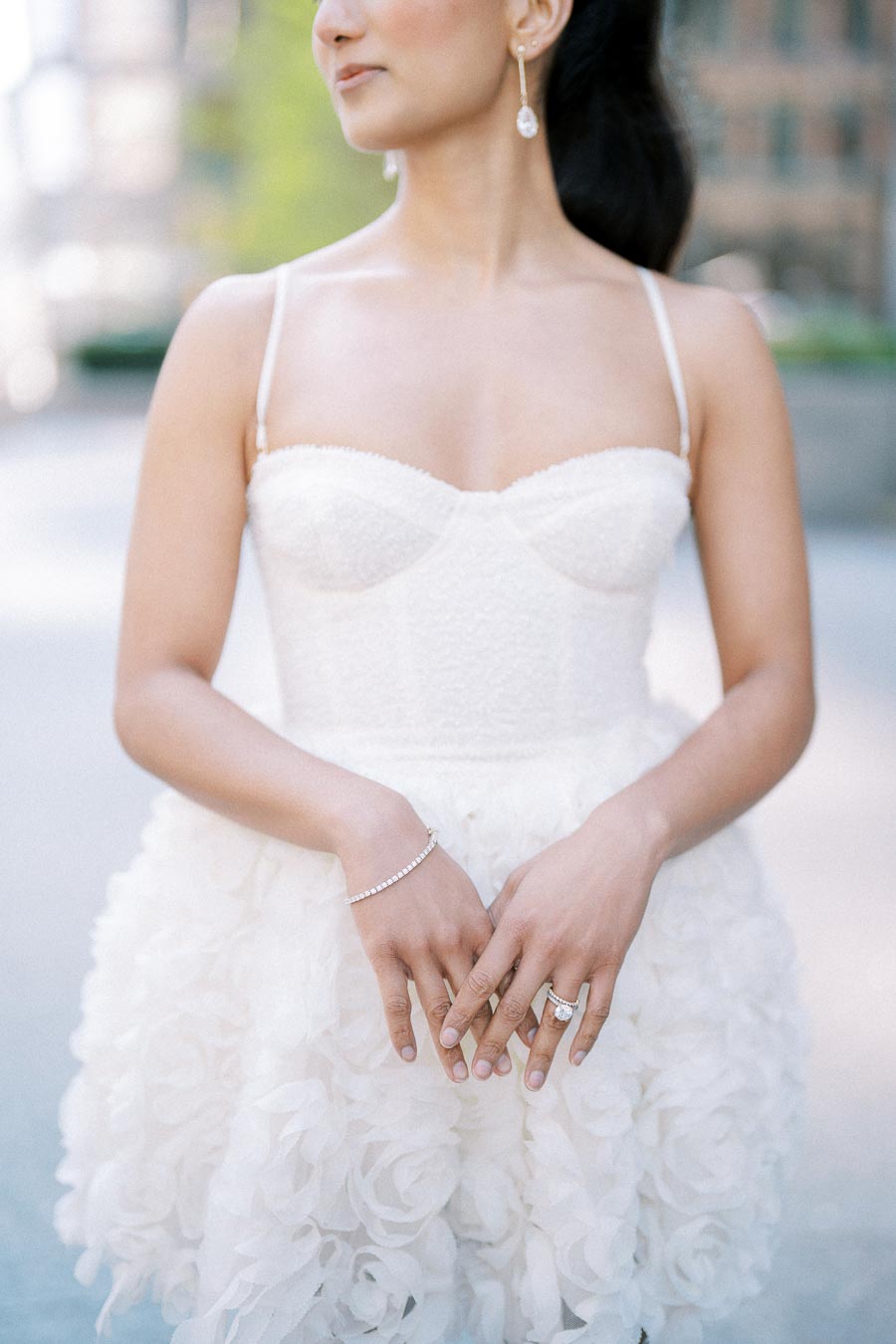 Elegant woman in a white wedding dress with floral details, showcasing diamond jewelry in an outdoor setting.