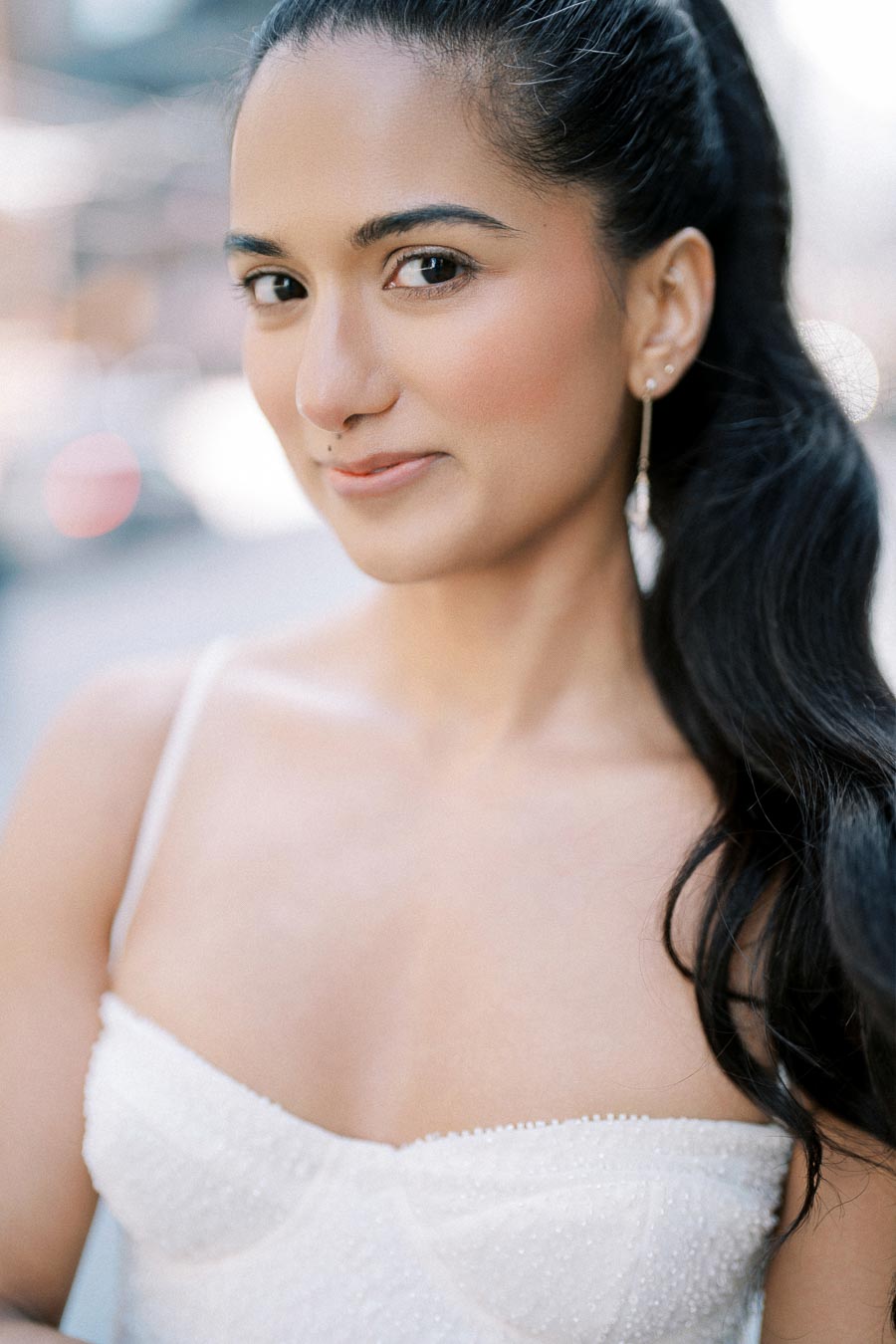 A woman with long black hair and a bright smile wearing a white strapless top, standing outdoors with a blurred urban background.