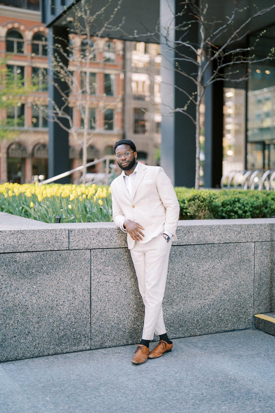 A stylish man in a light beige suit leans casually against a granite wall in an urban setting, surrounded by blooming yellow flowers and modern architecture.
