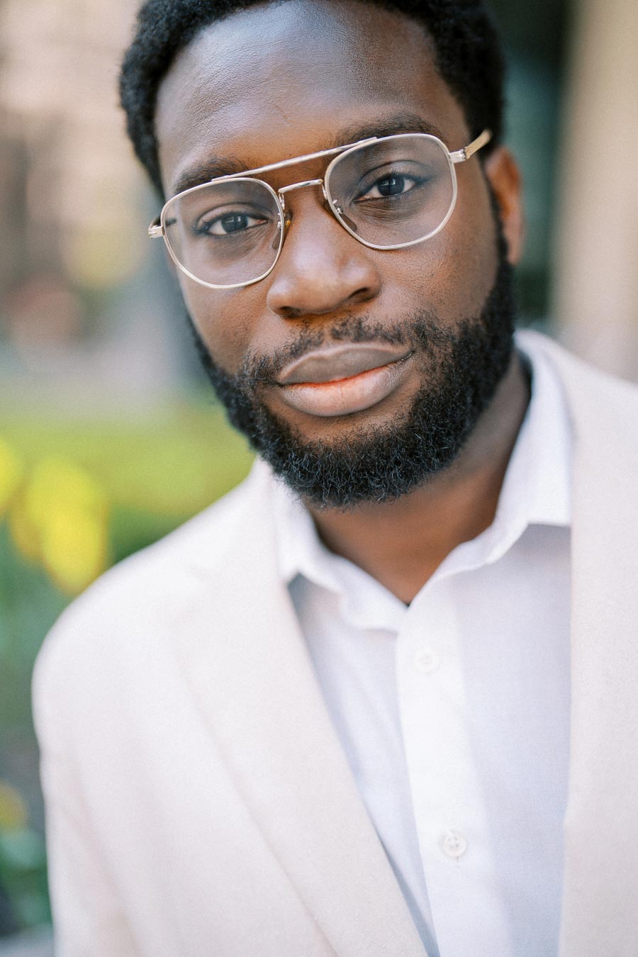 Portrait of a man with glasses and a beard wearing a light-colored suit jacket and white shirt, standing outdoors.