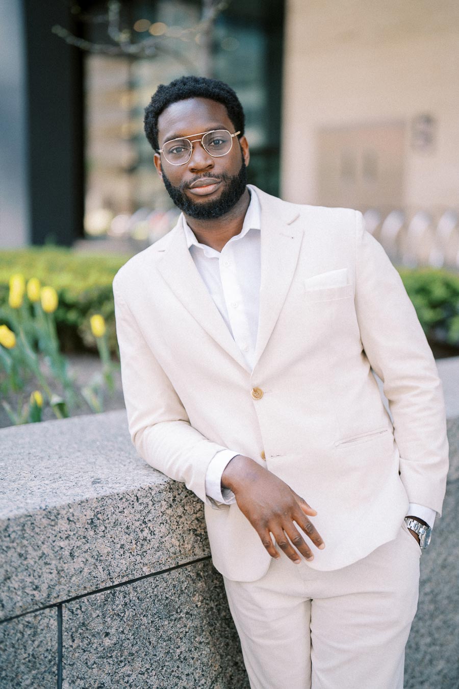 A well-dressed man in a light beige suit and glasses standing outdoors, leaning against a stone wall with yellow flowers in the background.