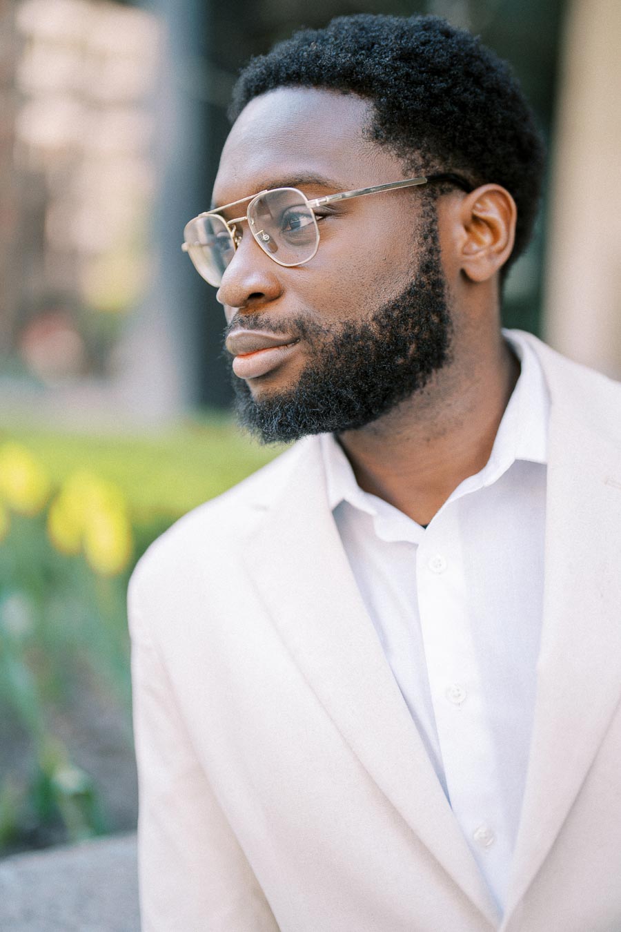 A man wearing glasses and a white suit looks thoughtfully into the distance outdoors, with greenery in the background.