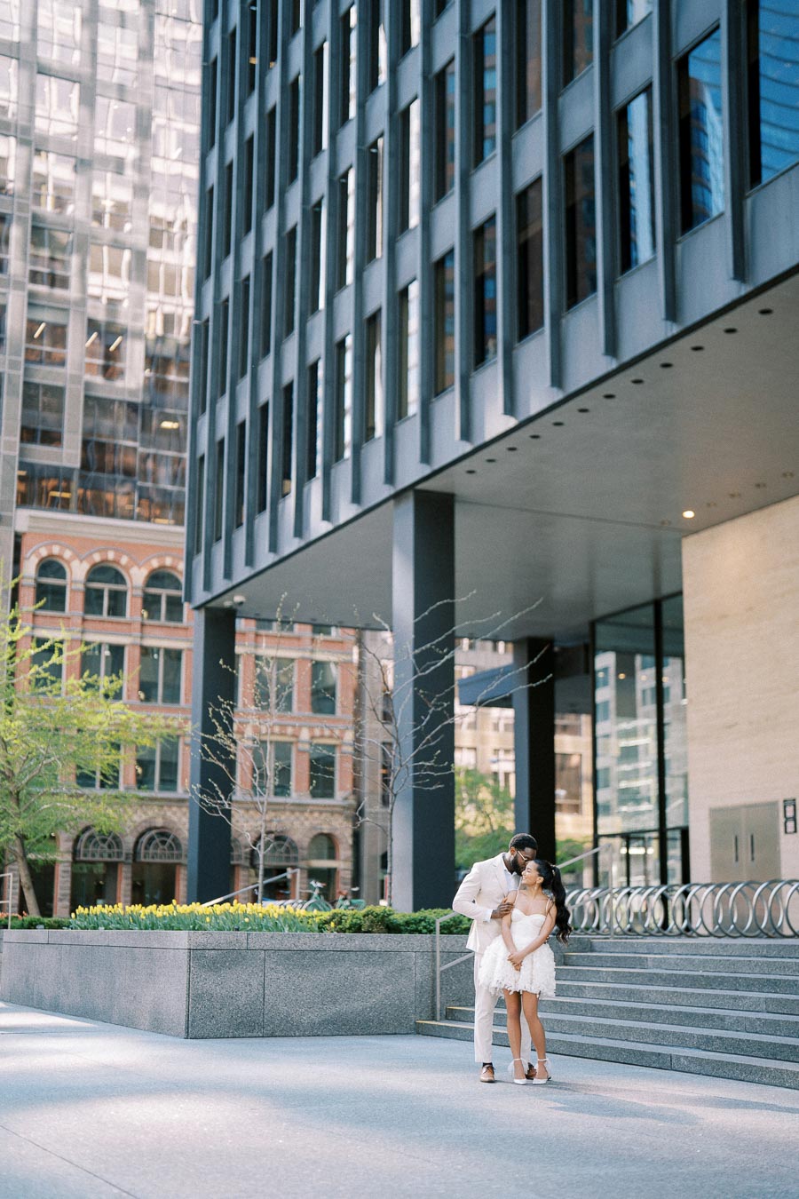 Couple embracing in elegant attire outside a modern city office building with large windows, surrounded by urban architecture and greenery.