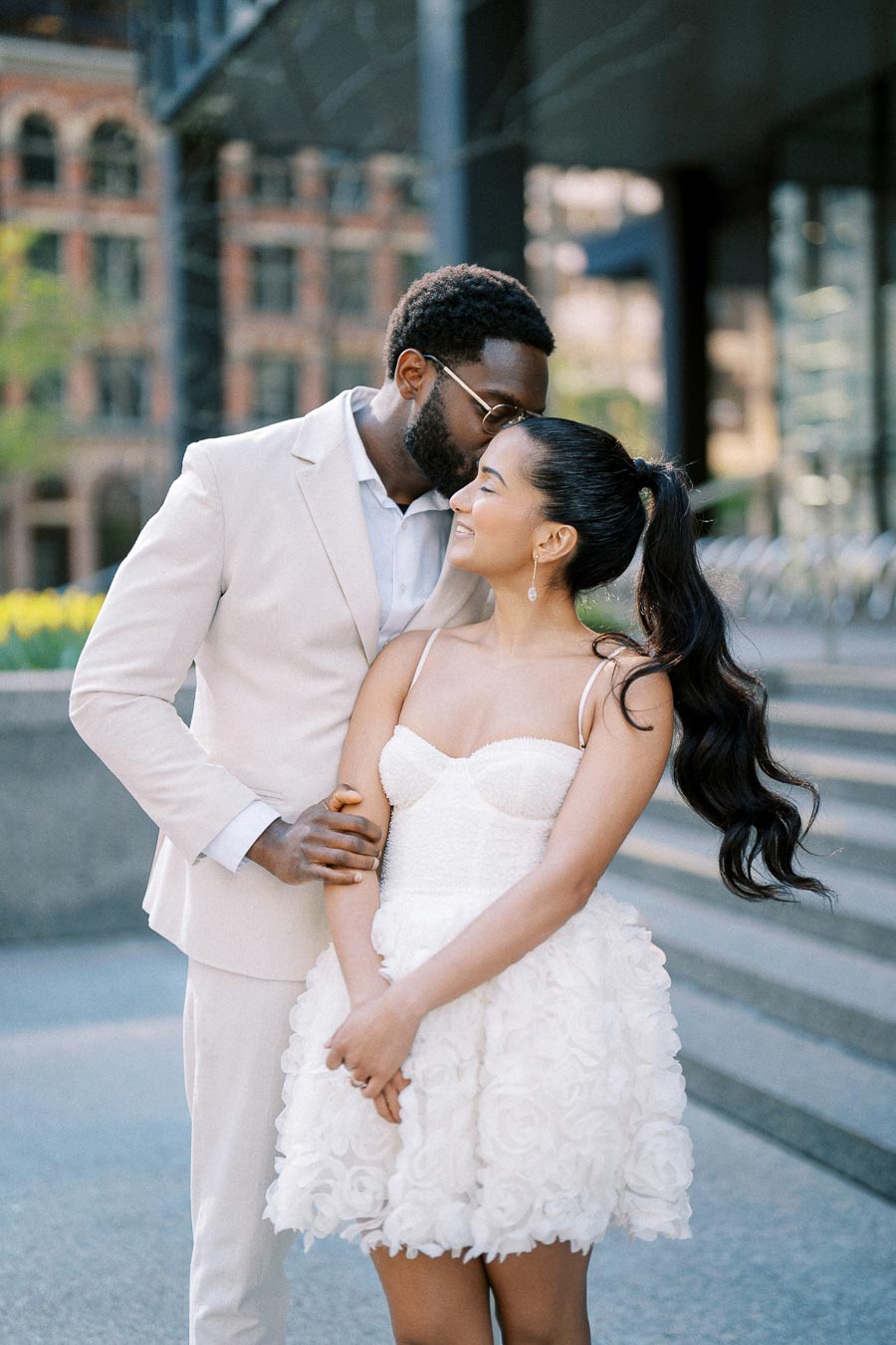A couple embracing in elegant attire, with the man in a light suit and the woman in a white floral dress, sharing a romantic moment outdoors in an urban setting.