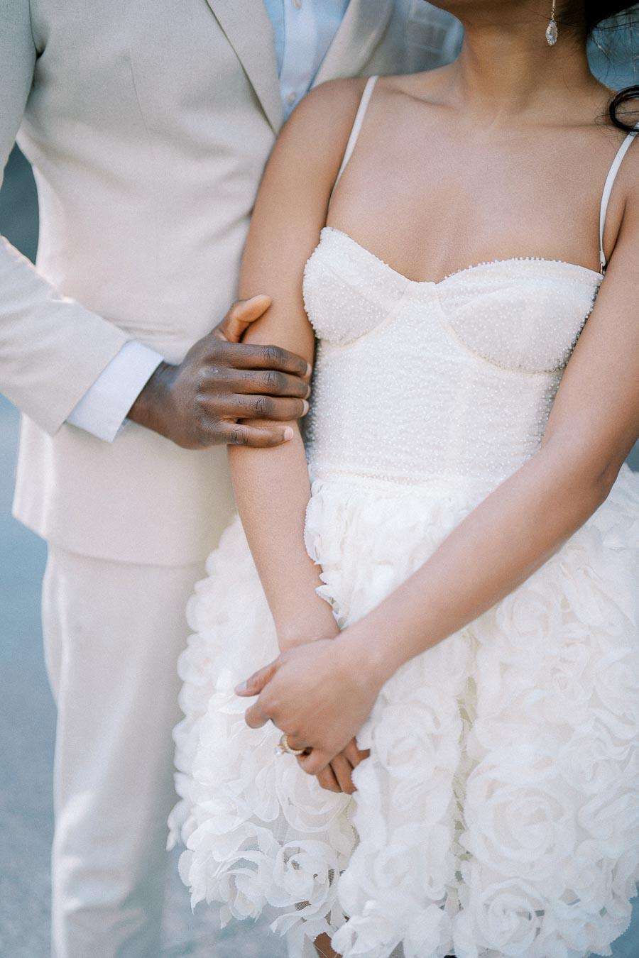 A couple in elegant wedding attire, with the bride wearing a detailed white floral dress and the groom in a light suit, sharing an intimate embrace.