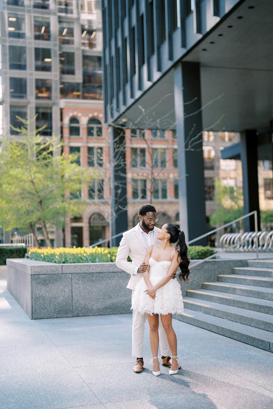 Happy couple embracing in an urban setting with modern architecture and greenery, wearing elegant wedding attire.