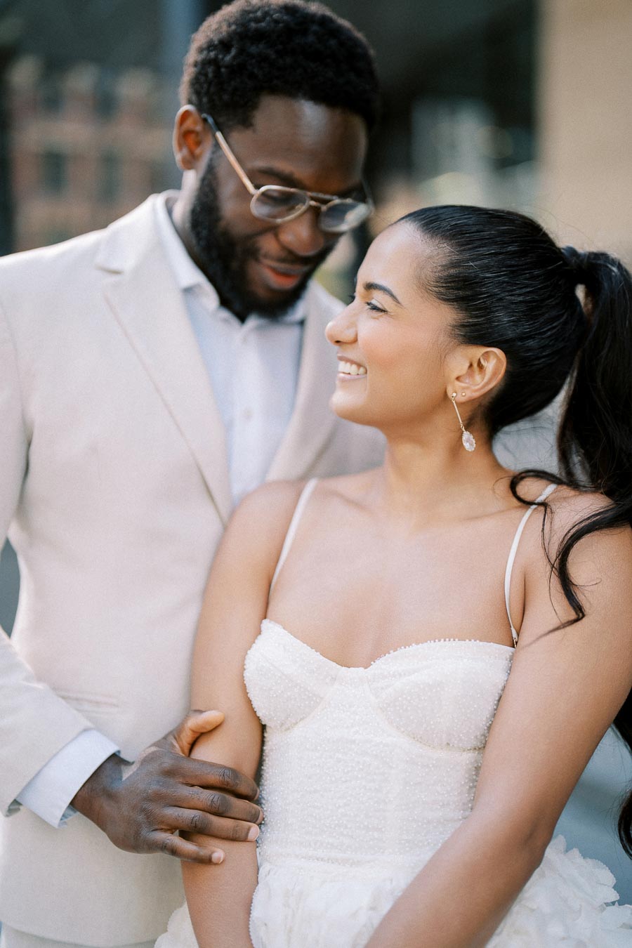 A joyful couple embraces on their wedding day, with the bride smiling in a white dress and the groom in a light suit, under soft natural lighting.