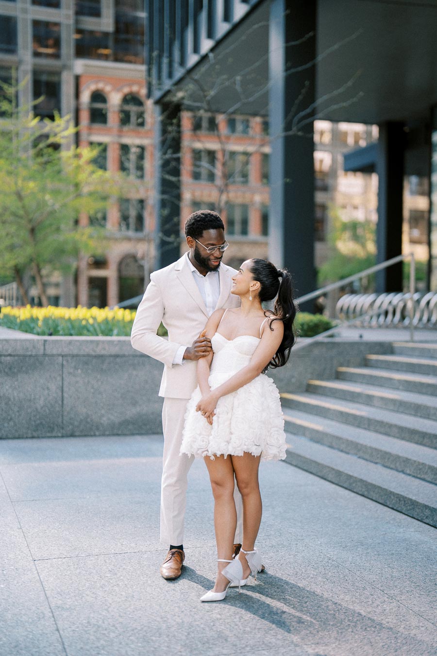 A couple in elegant attire shares an affectionate moment outdoors. The man is wearing a light suit, and the woman is in a short white dress with floral details. They stand on a modern city street with steps and greenery in the background, exuding romance and happiness.