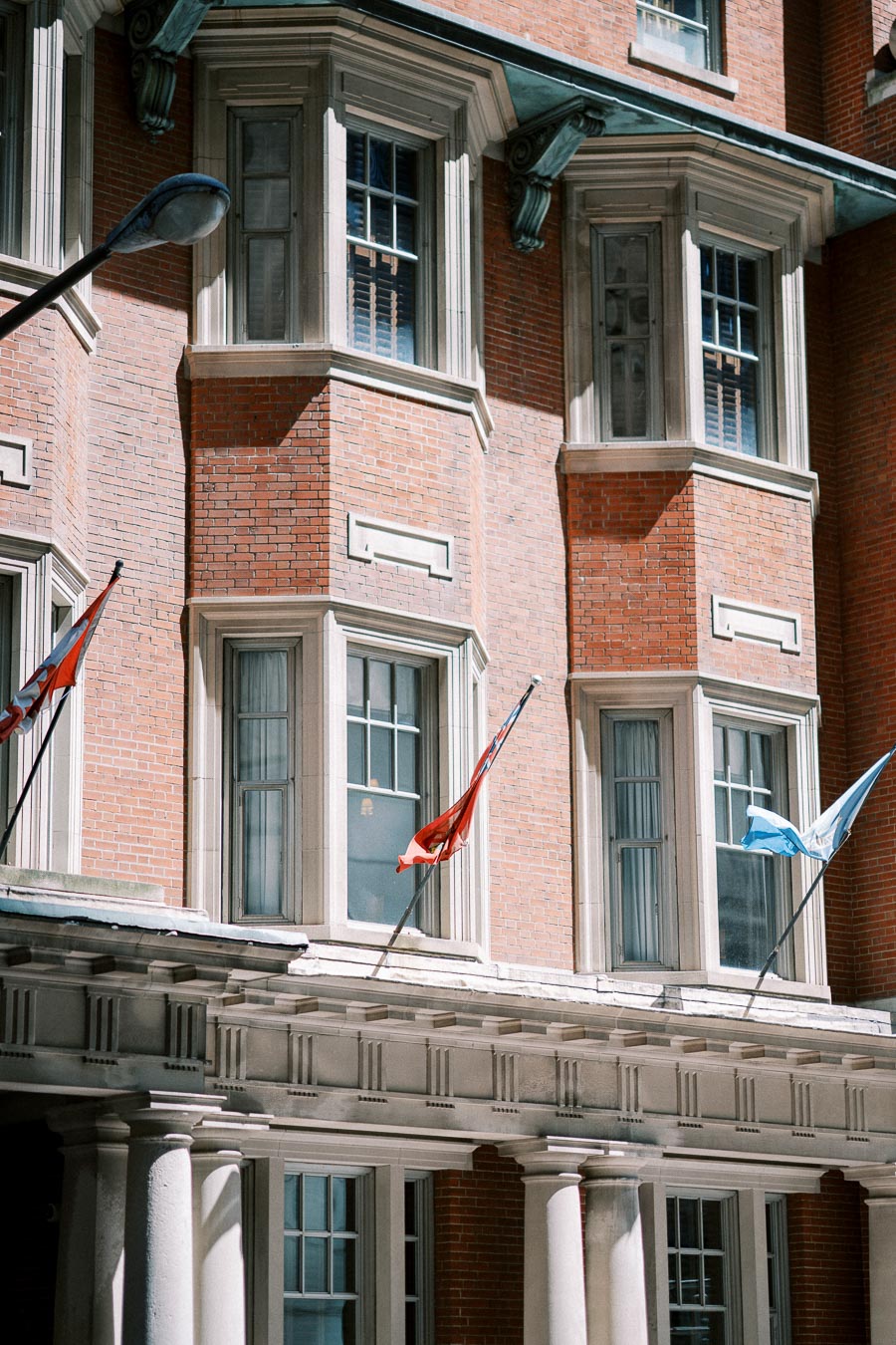 Historic red brick building facade with bay windows and international flags, showcasing classic architectural details and columns, in bright daylight.