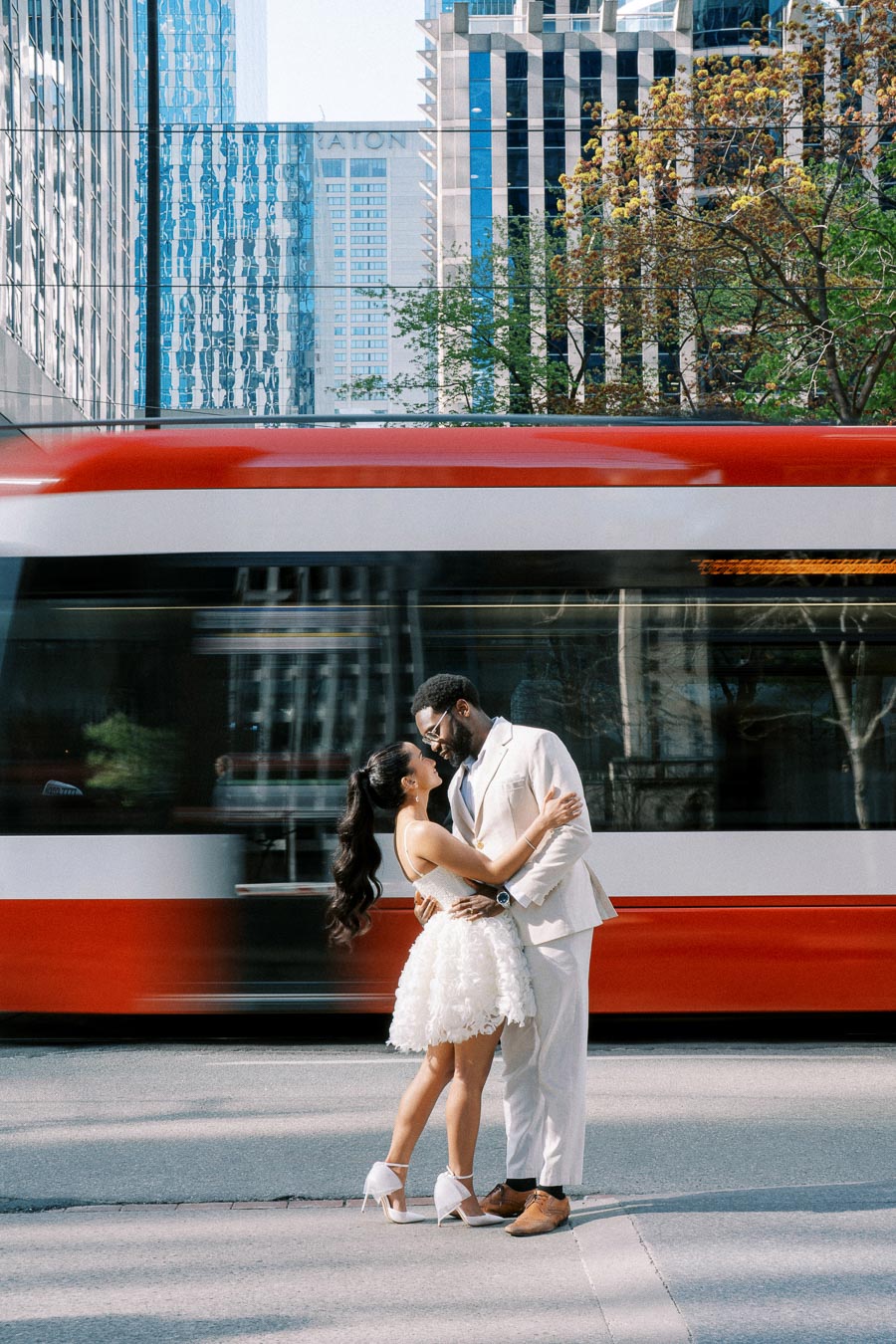 Engaged couple embracing on city street with blurred red and white tram passing by, surrounded by modern skyscrapers and greenery.
