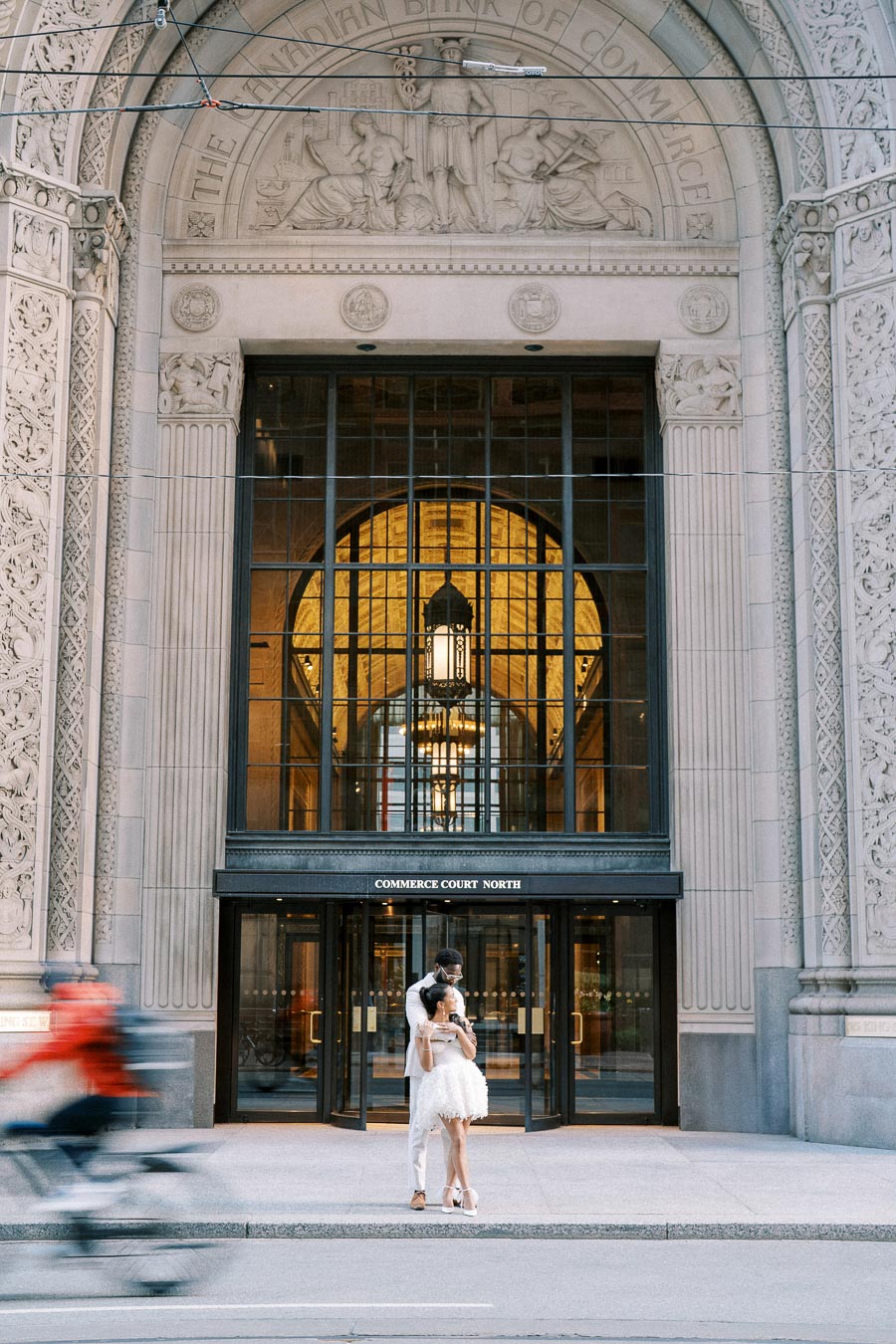 A couple embracing in front of the ornate entrance of Commerce Court North in Toronto, with a cyclist passing by in the foreground.