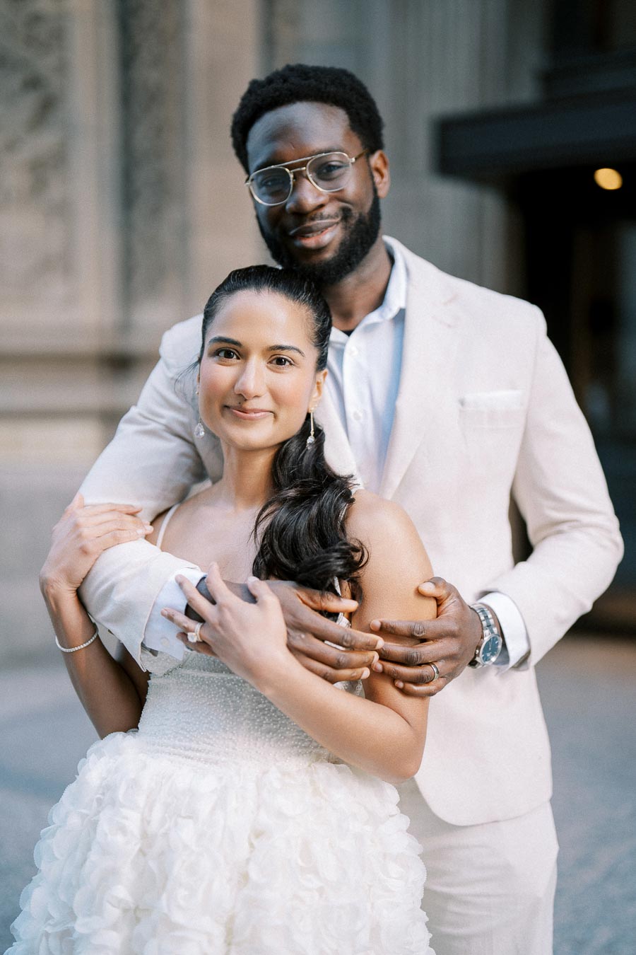A couple posing together in elegant attire, with the man embracing the woman from behind. The woman is wearing a white dress with a ruffled skirt, while the man is dressed in a light-colored suit. They are smiling softly, standing in an outdoor setting with architectural details in the background.