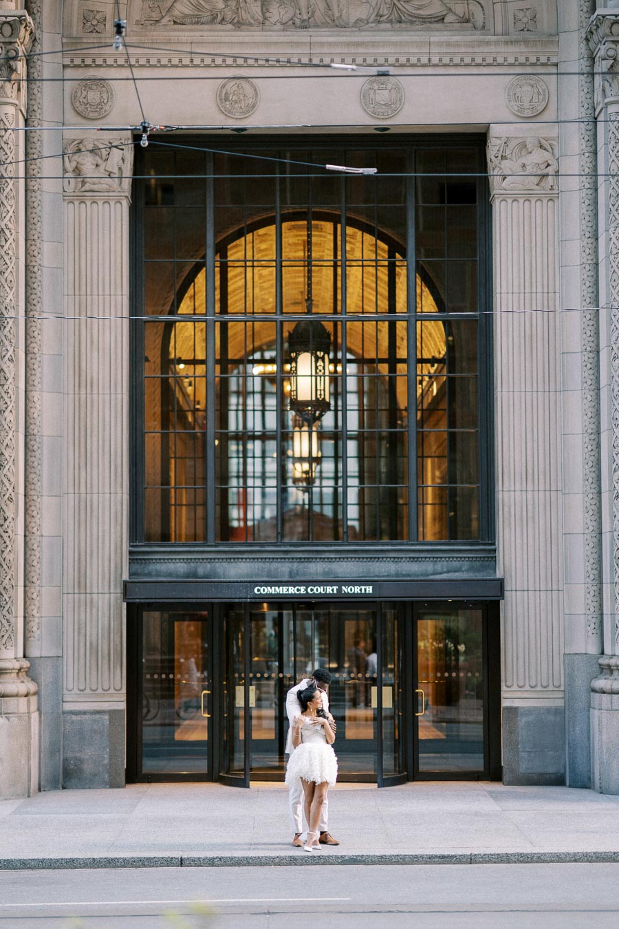Couple embracing in front of the historic entrance of Commerce Court North building, showcasing elegant architecture with grand arched windows and intricate carvings in a downtown setting.