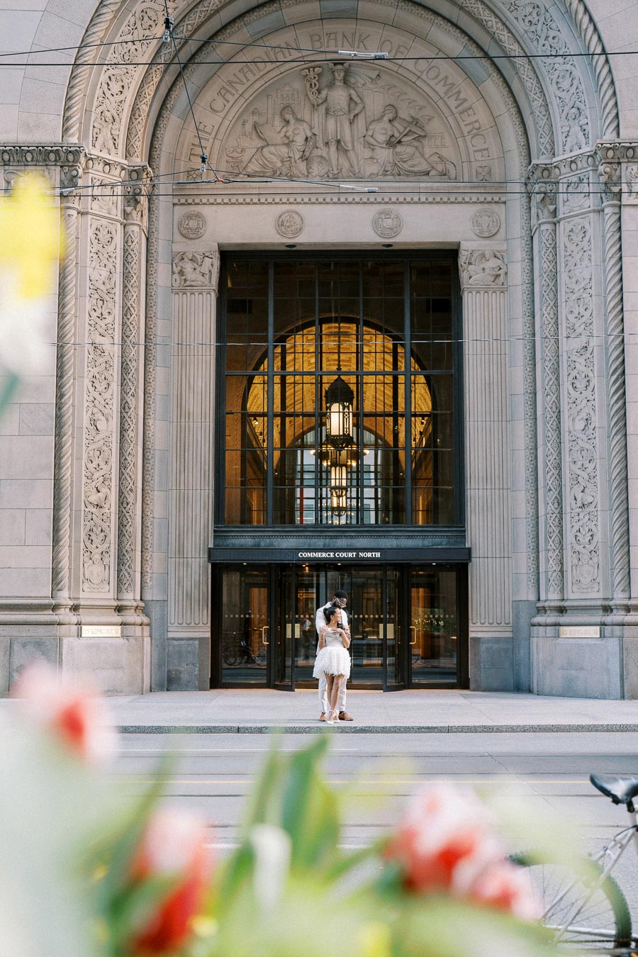 Engaged couple embracing in front of ornate entrance at Commerce Court North, framed by vibrant flowers in the foreground