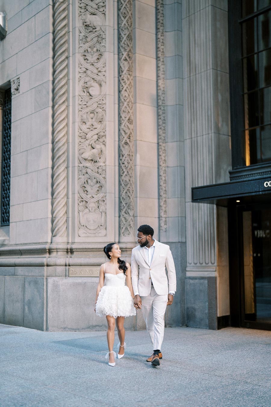 Couple walking hand in hand, dressed elegantly in white attire, in front of a beautifully ornate building.