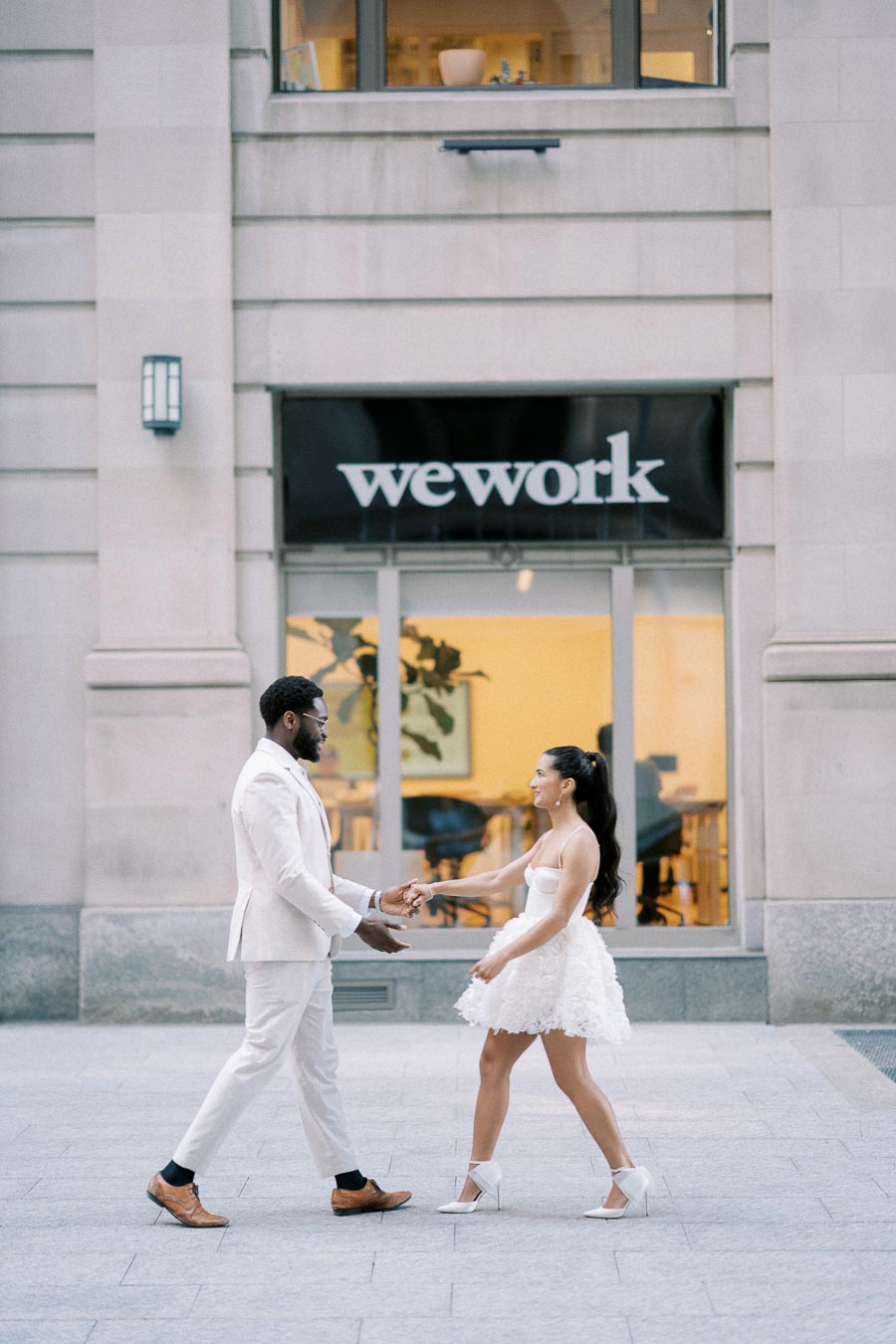 A couple dressed in elegant white outfits dancing outside a WeWork office building, capturing a joyful and romantic moment.