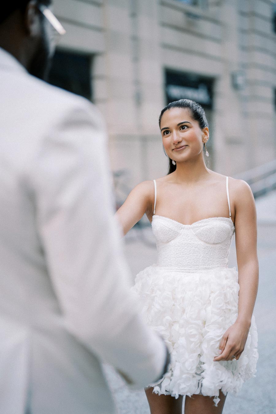 A woman in a white dress smiling outdoors, extending her hand towards a person in a white suit, with a city building in the background.