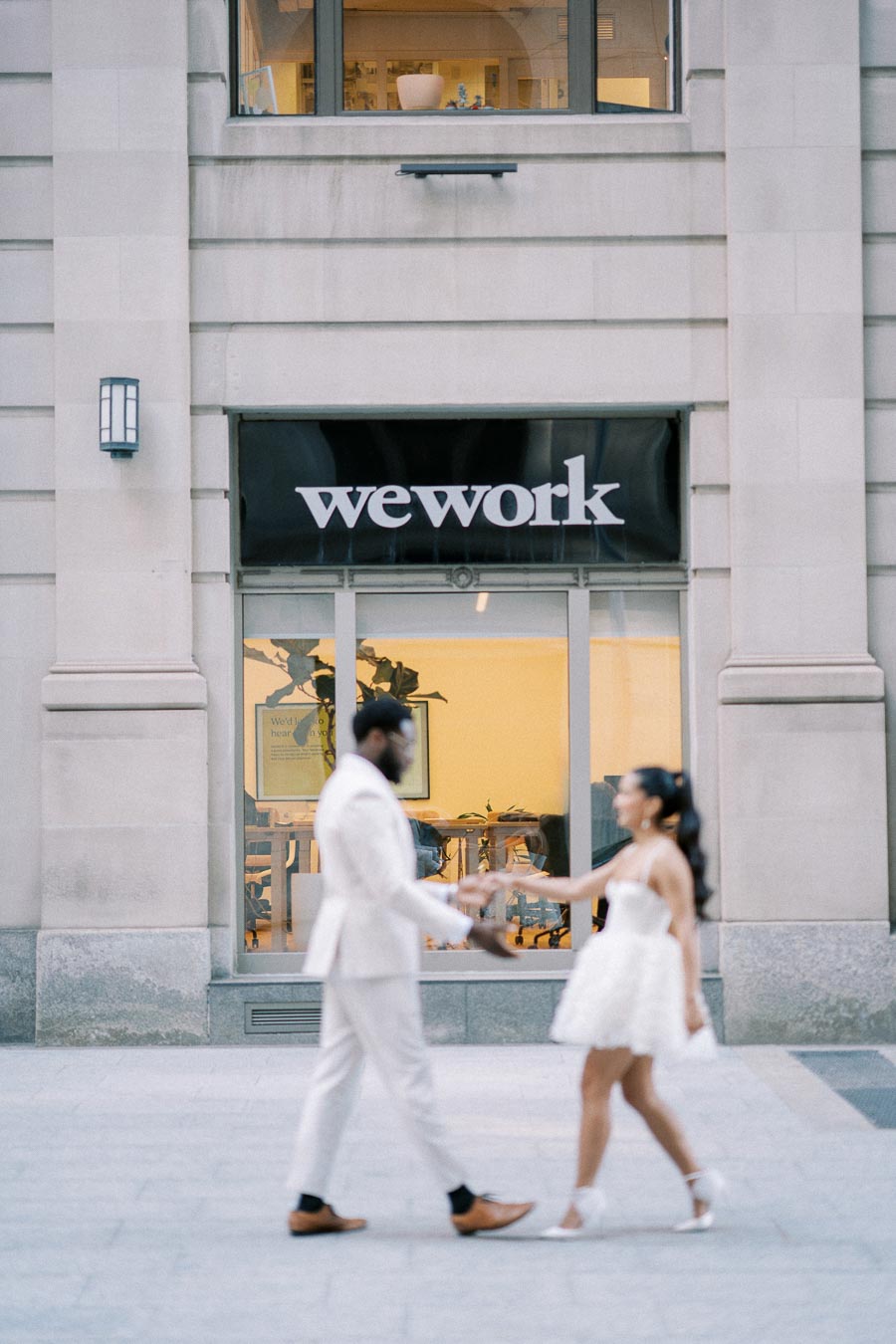 A couple in elegant white attire holds hands while standing outside a WeWork office building, symbolizing connection and modern workspace collaboration.