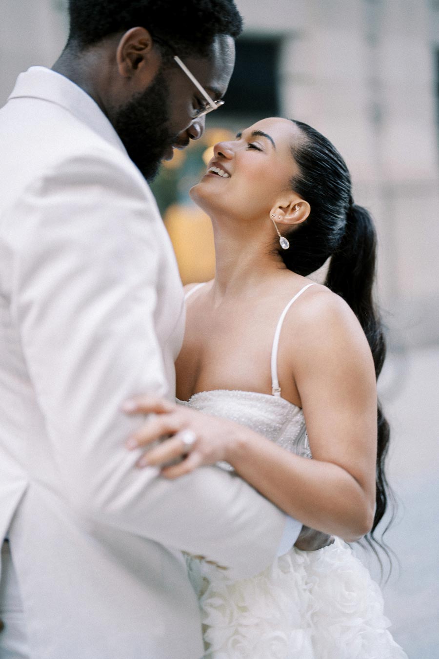 A bride and groom embracing and smiling at each other in an elegant outdoor setting, with the bride wearing a strapless wedding gown adorned with floral details and the groom in a light-colored suit, creating a romantic and joyful wedding moment.
