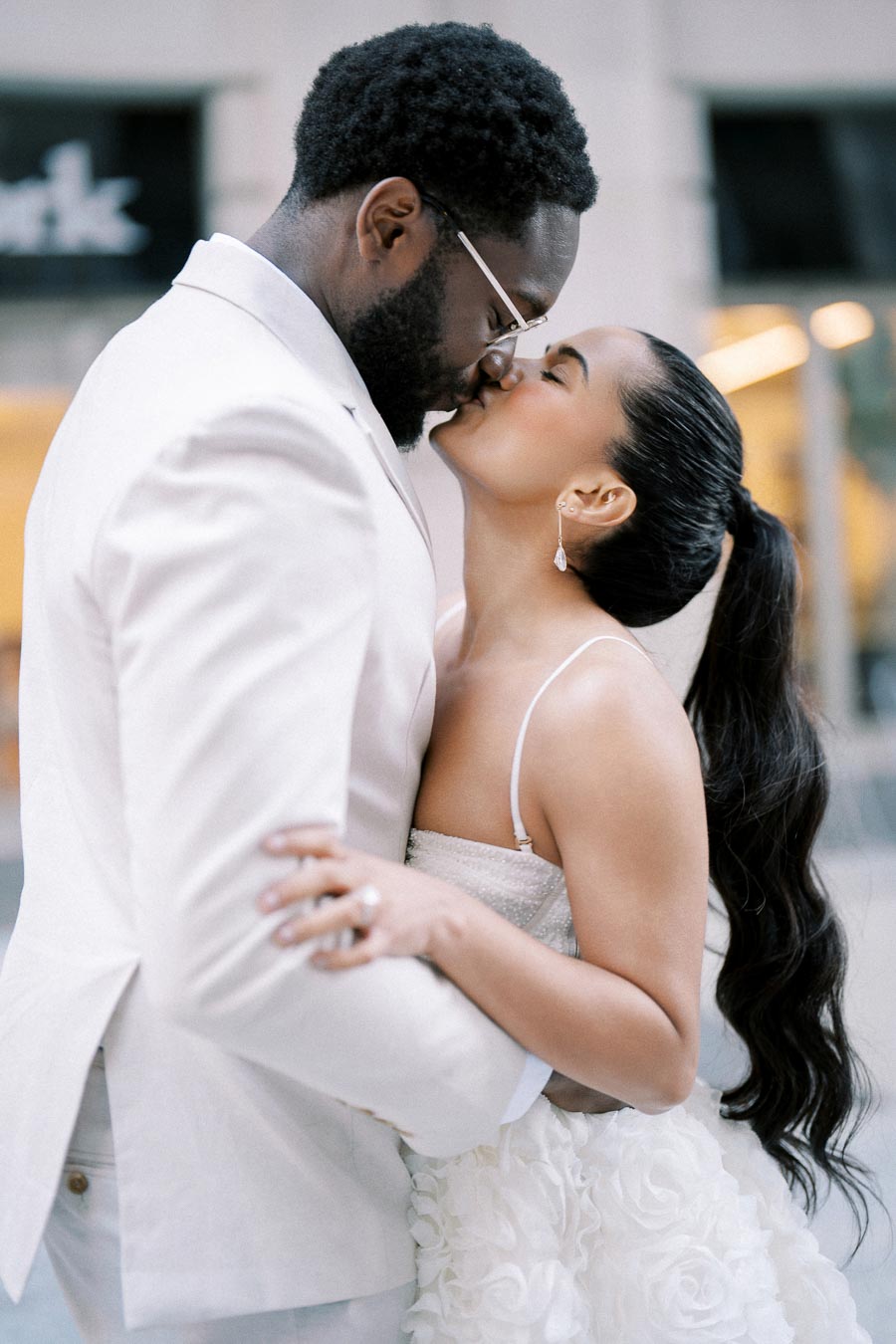 Romantic couple embracing and kissing outdoors, wearing elegant white attire, with a soft focus cityscape background.
