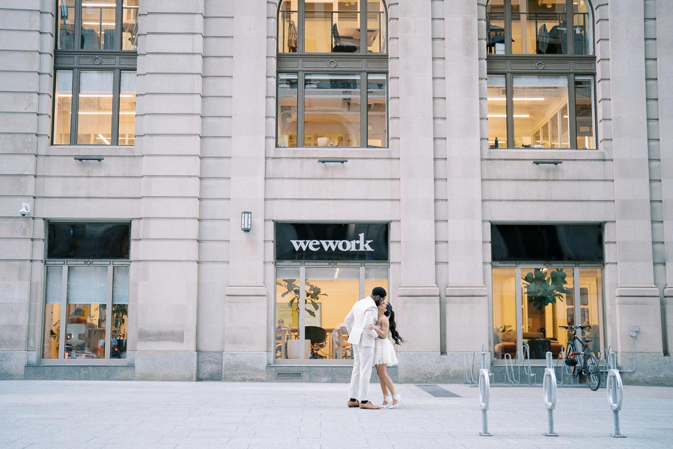 A couple embraces romantically in front of a WeWork building with large windows, showcasing a modern office interior, against a backdrop of urban architecture.