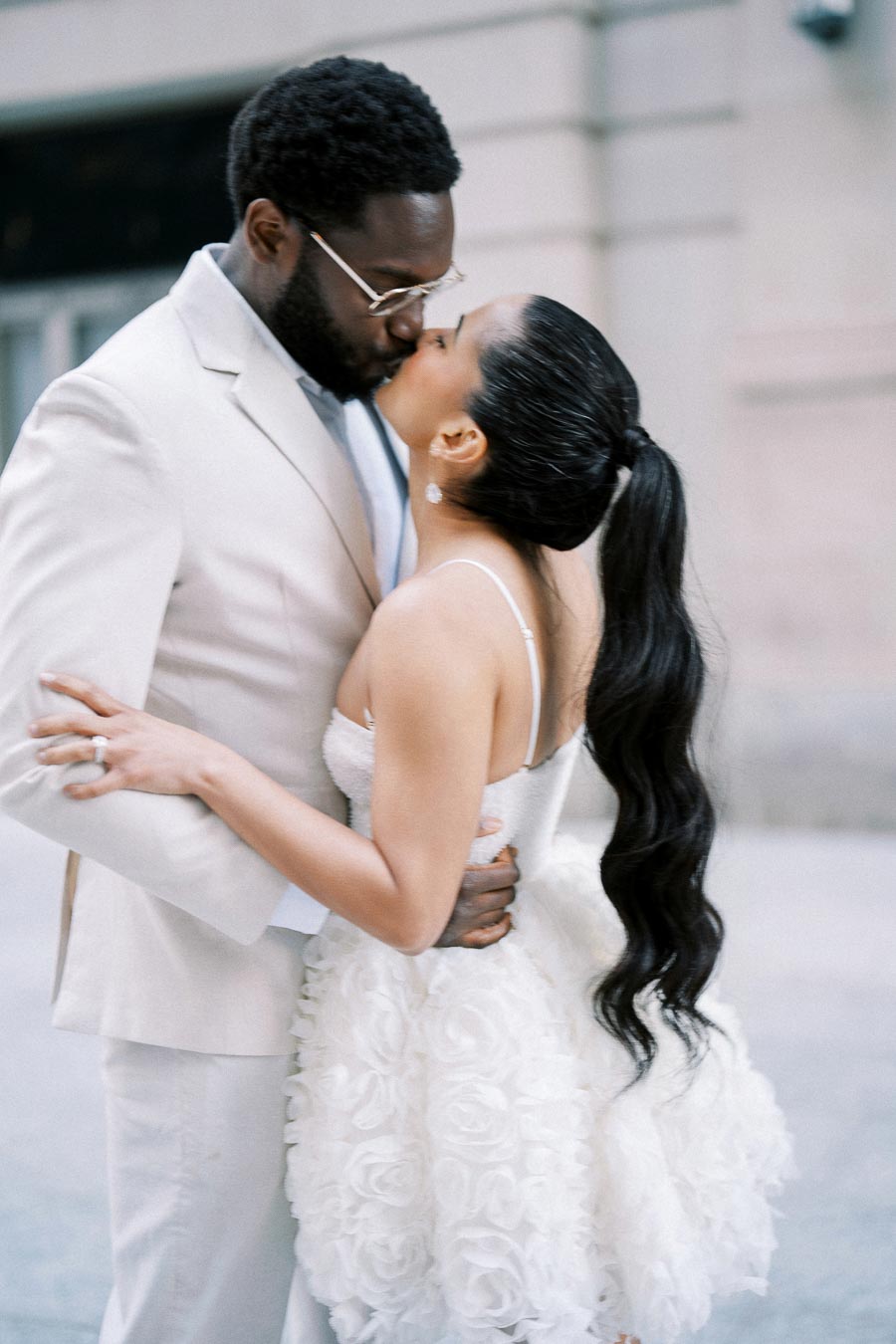 A couple in elegant attire shares a romantic kiss outdoors, with the woman in a white floral dress and the man in a light-colored suit, against a blurred urban background.