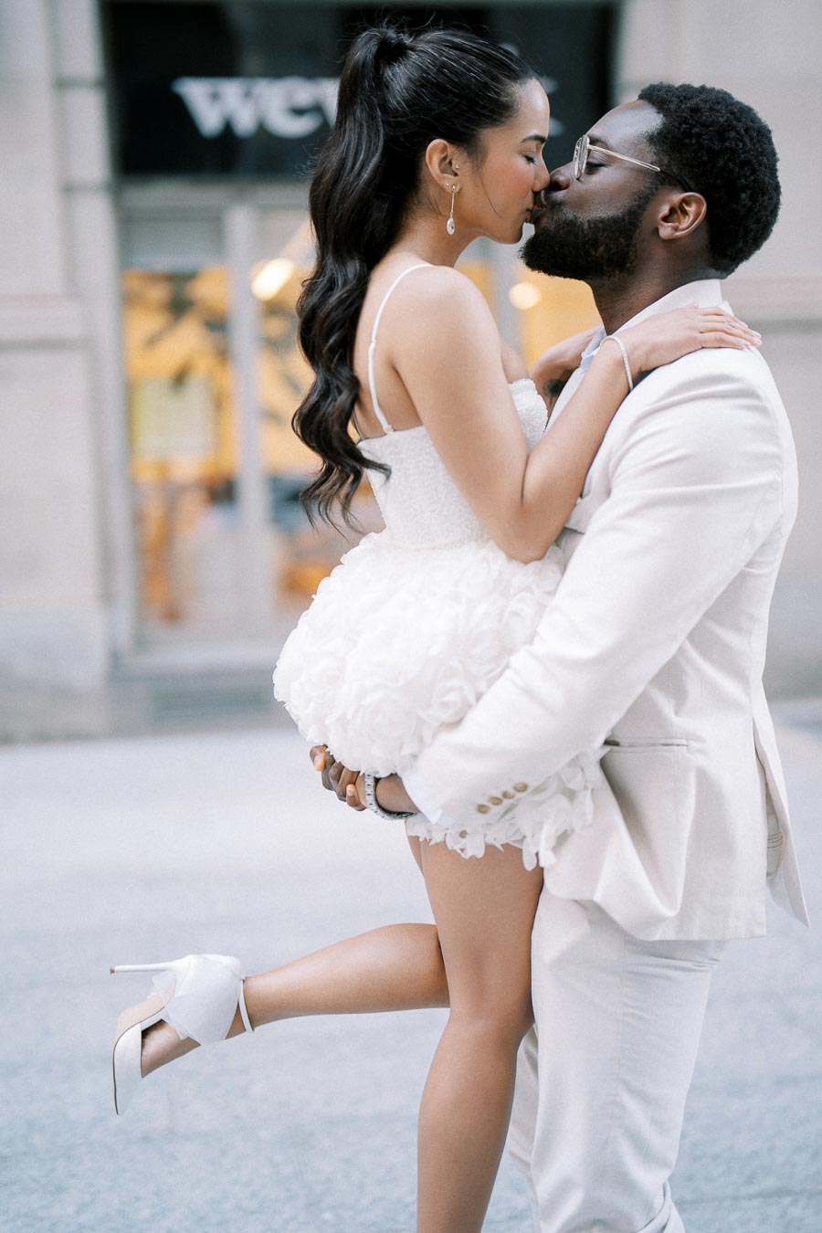 Young couple in elegant white attire sharing a romantic kiss on a city street.