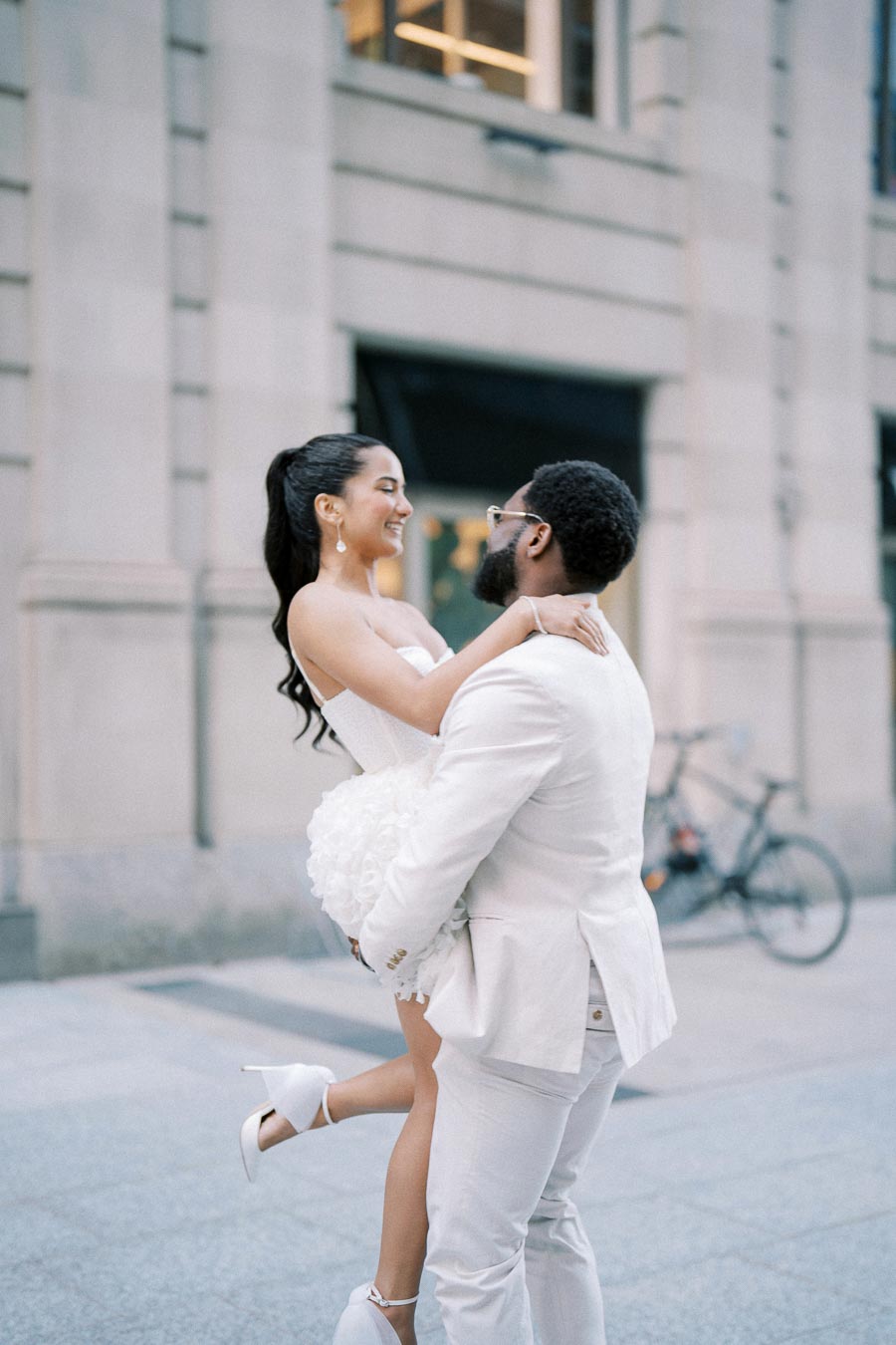 A joyful couple in wedding attire, with the groom lifting the bride against an urban backdrop, embodying love and celebration on their special day.