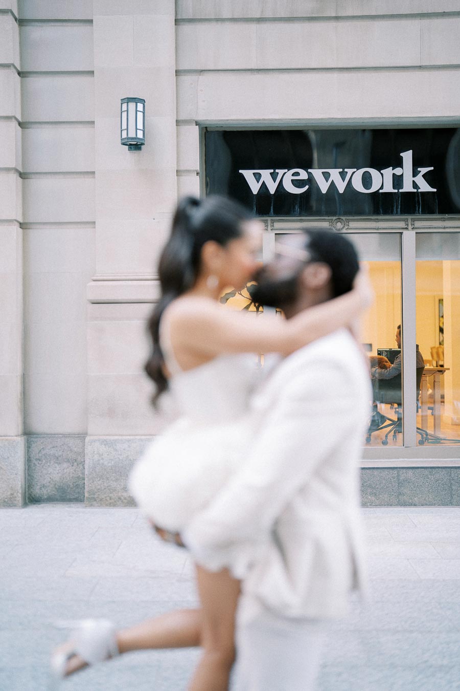 A couple embracing in front of a WeWork building, with both dressed in elegant white outfits, capturing a romantic moment in an urban setting.