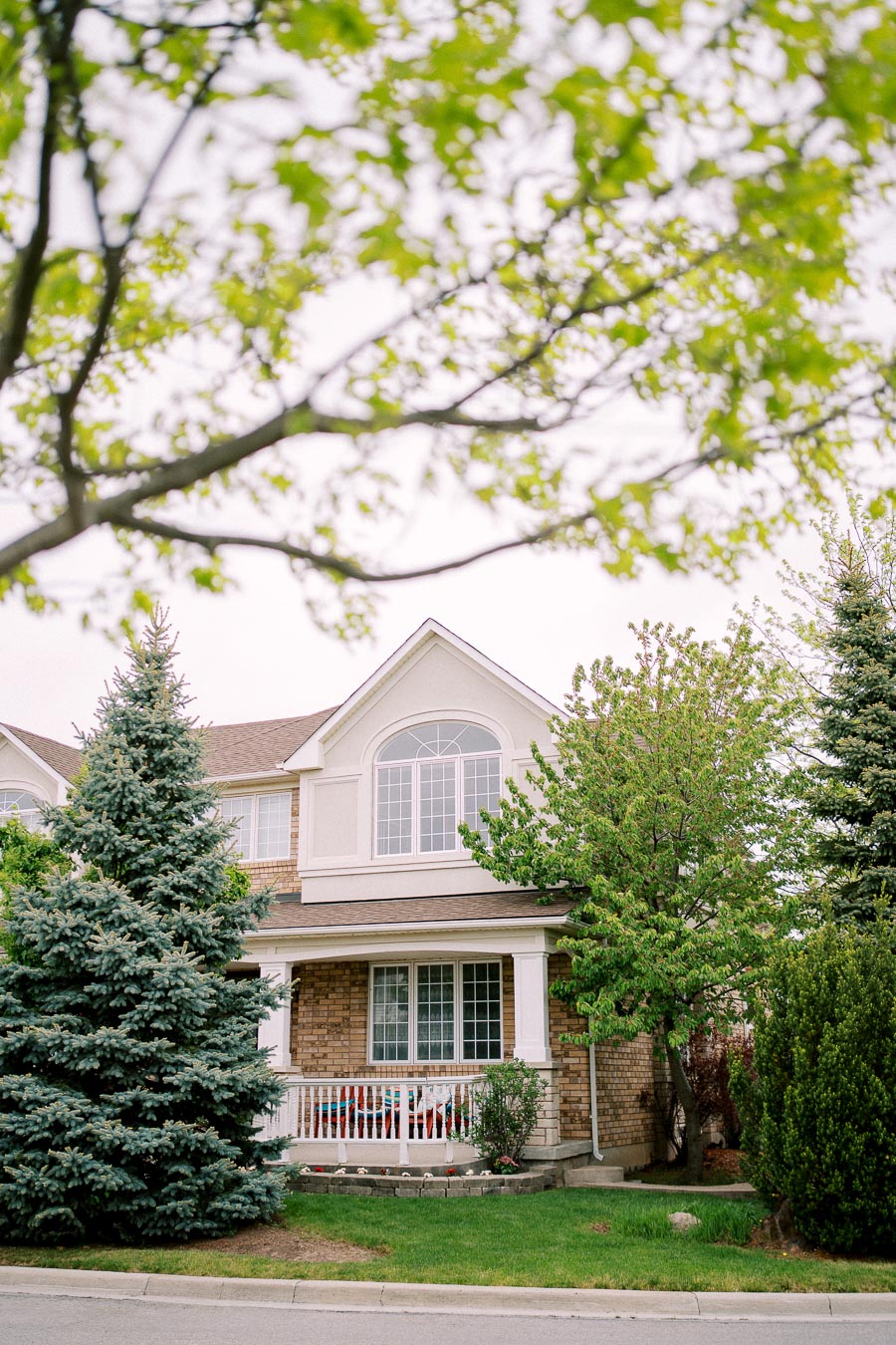 Charming suburban house with brick facade and gabled roof, surrounded by lush green trees and well-maintained lawn, viewed through foreground branches with fresh spring leaves.