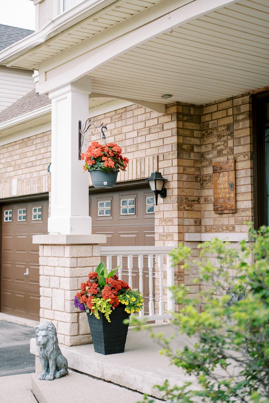 Alt text: Charming house porch with brick walls and decorative plants, featuring vibrant floral arrangements in a black planter and a hanging basket, with a stone lion statue on the step and brown garage doors in the background.
