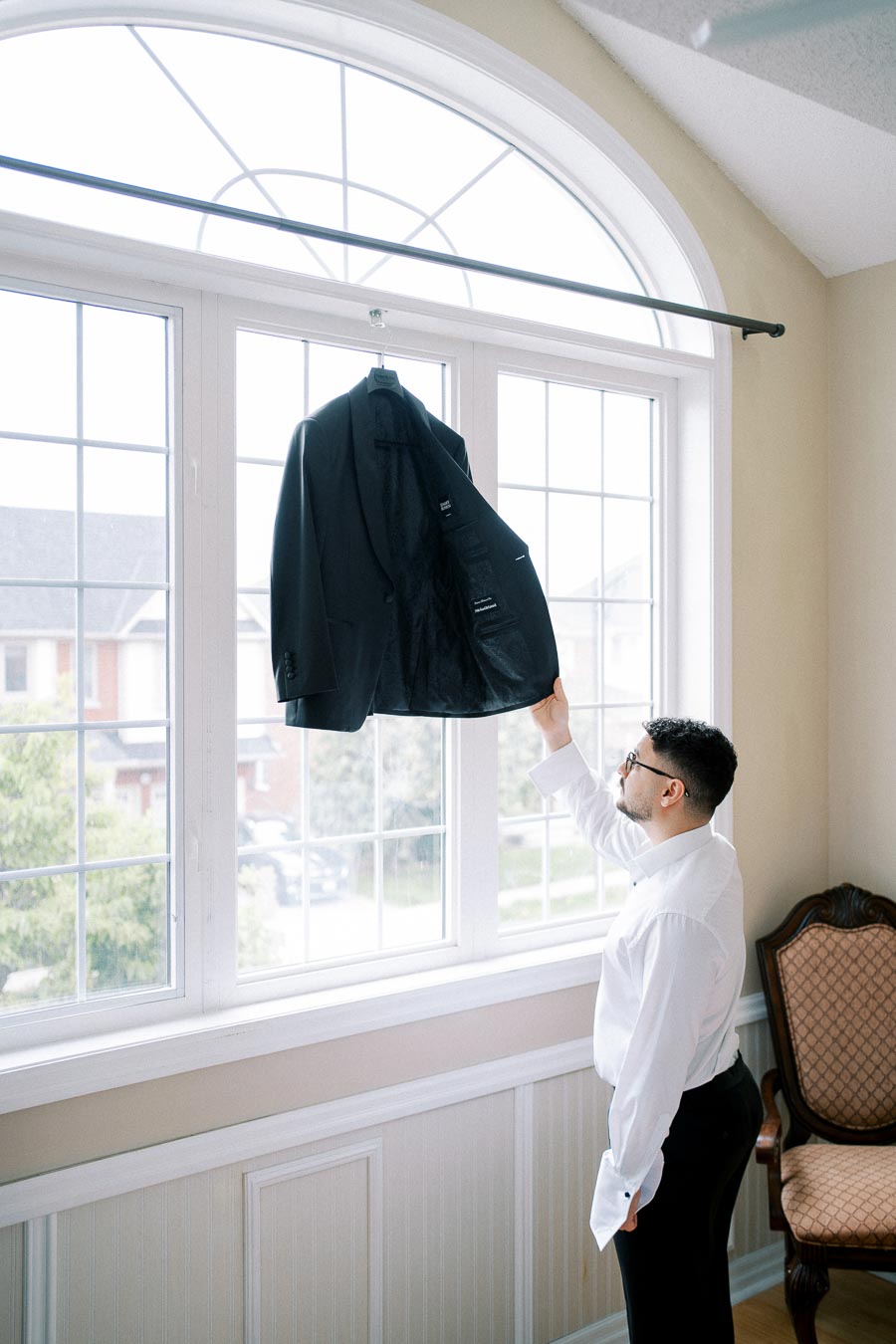 Man in white shirt adjusting a black suit jacket hanging on a window rod, in a bright room with large windows.