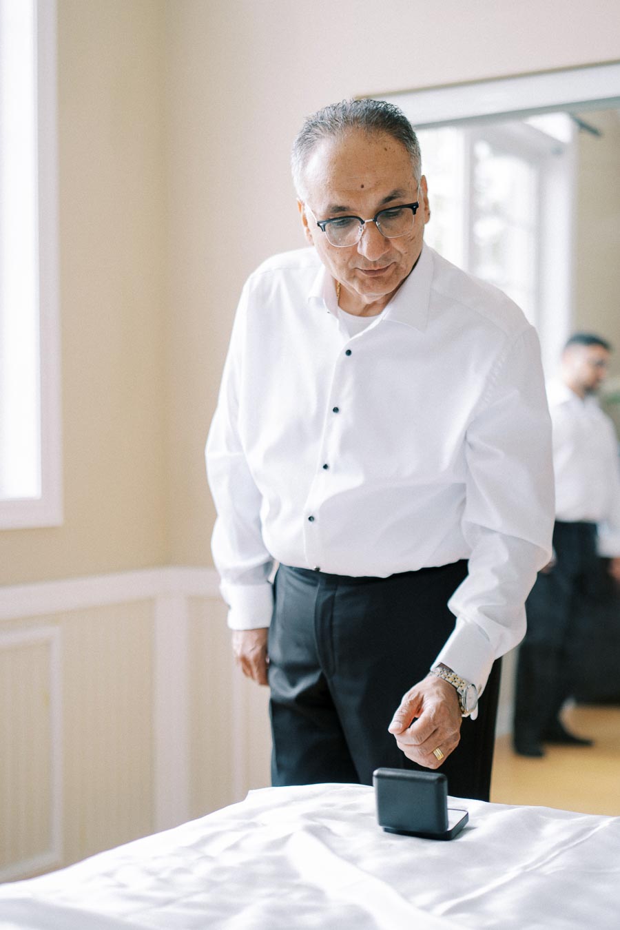 Elderly man in a white dress shirt and glasses, standing in a softly lit room, looking at a black jewelry box on a white tablecloth.