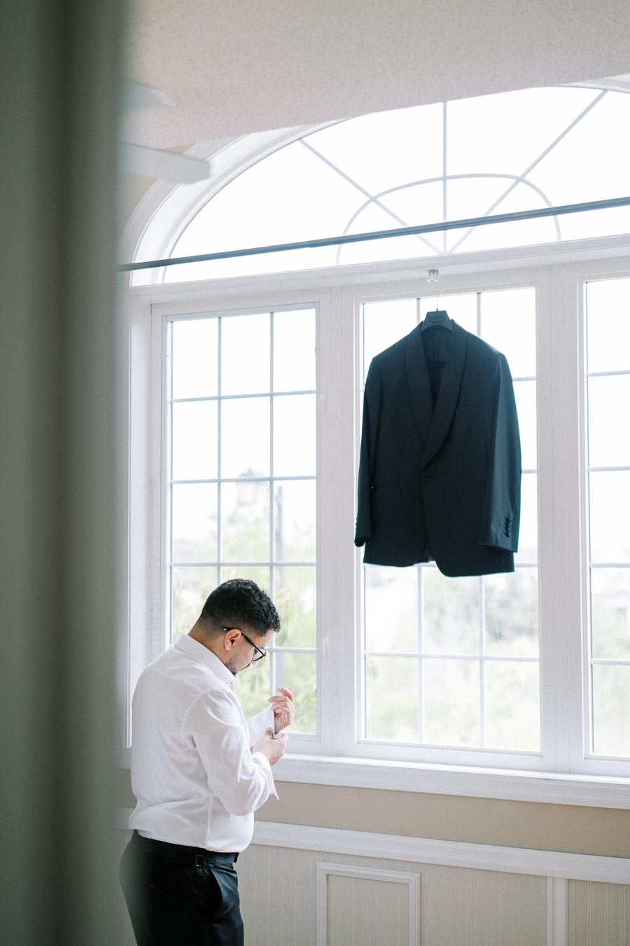A man in a white shirt adjusting his cufflinks next to a large window with a hanging black suit jacket, preparing for a formal event.