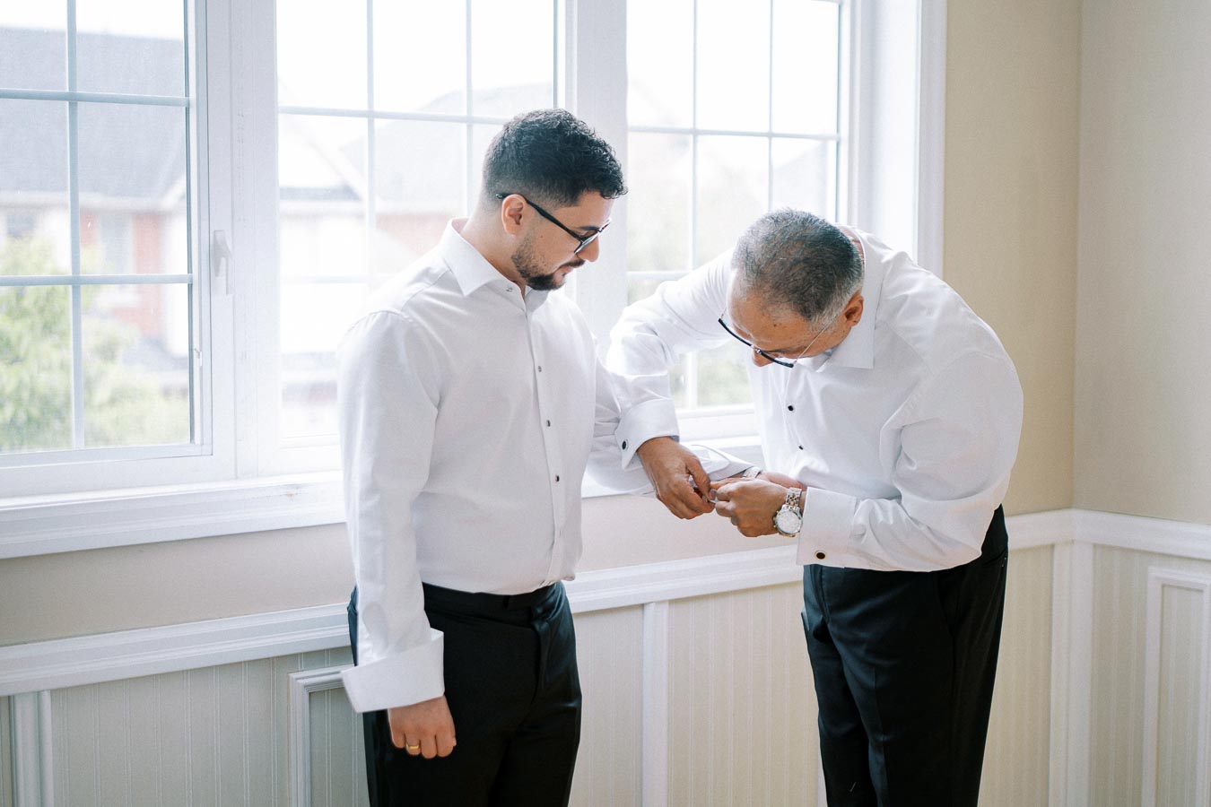 A young man in a white dress shirt is assisted by an older gentleman adjusting his cuffs in a bright room with large windows.